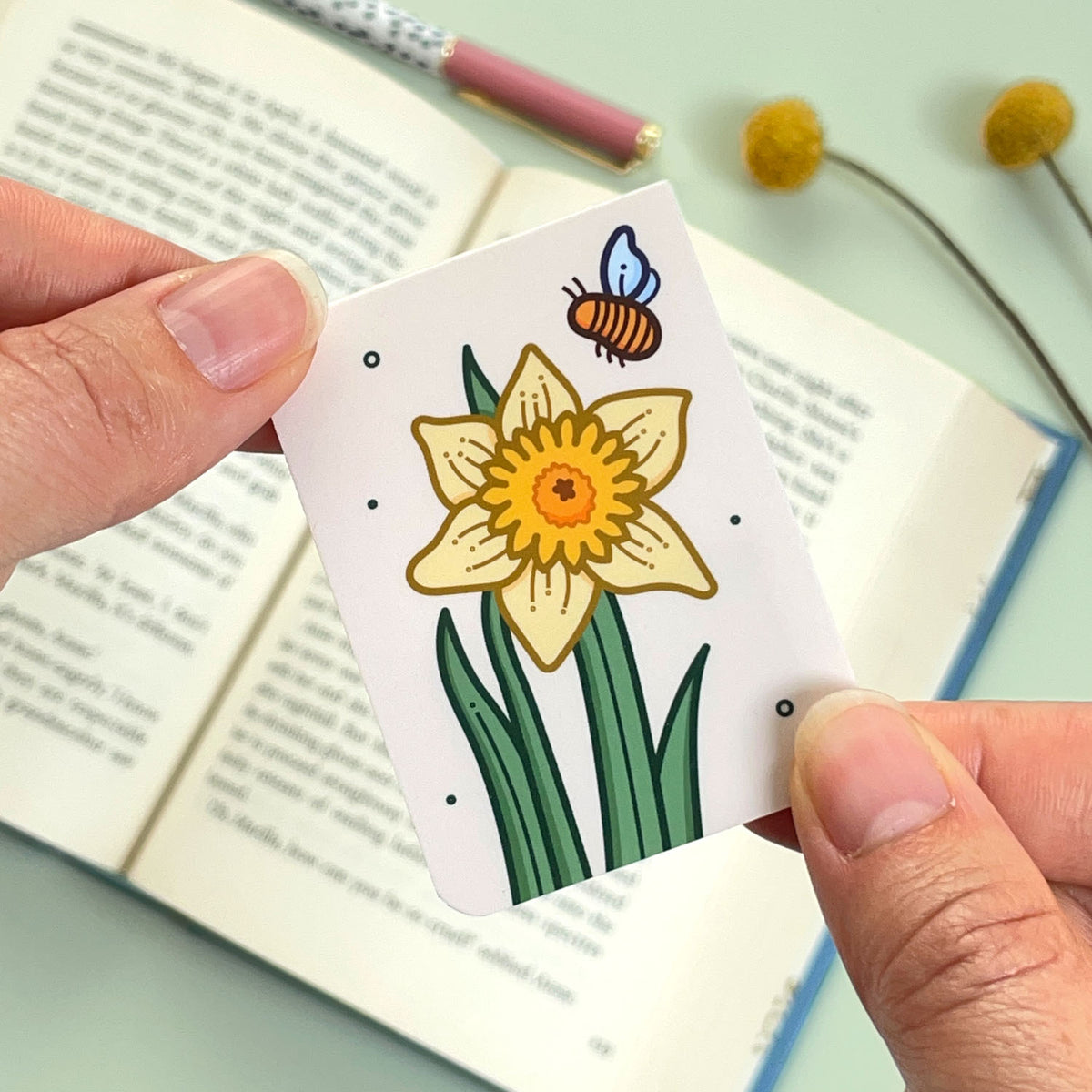The reverse side of the floral daffodil bookmark with a yellow flower and a bee held over an open book