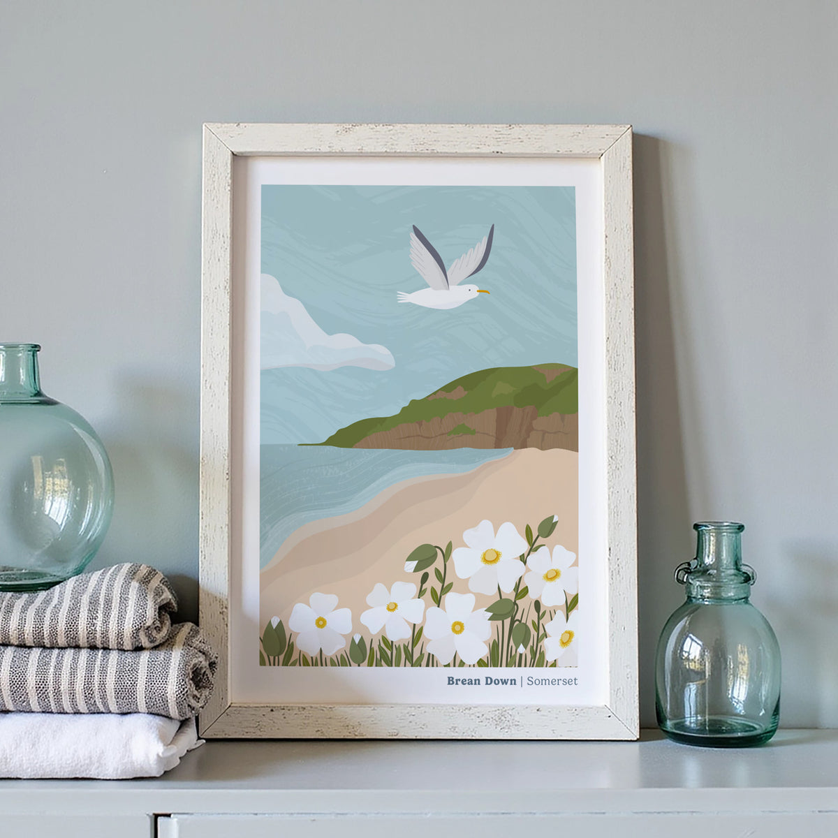 A framed art print featuring an illustrated coastal landscape of Brean Down in Somerset with a flying seagull, calm beach, green cliffs, and white flowers, displayed on a shelf with folded towels and glass bottles.