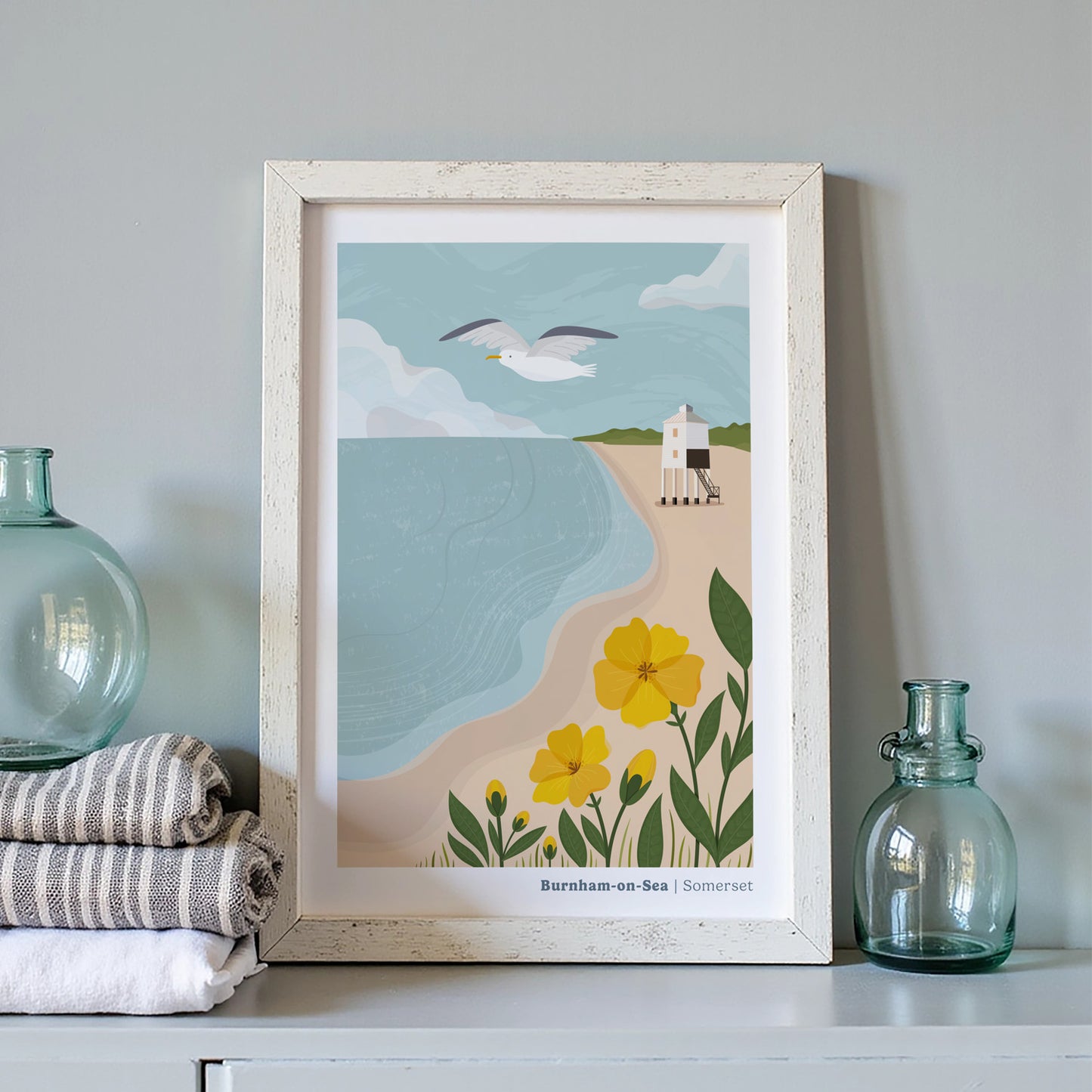 A coastal art print of the Low Lighthouse at Burnham-on-Sea inside of a white picture frame next to two blue bottles and a pile of linen cloths.