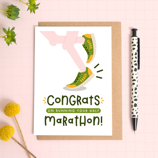 A half marathon congratulations card photographed on top of a kraft brown envelope set on a pink background surrounded by foliage and a pen for scale. The card reads 'congrats on running your half marathon' and features a pair of pale pink legs.