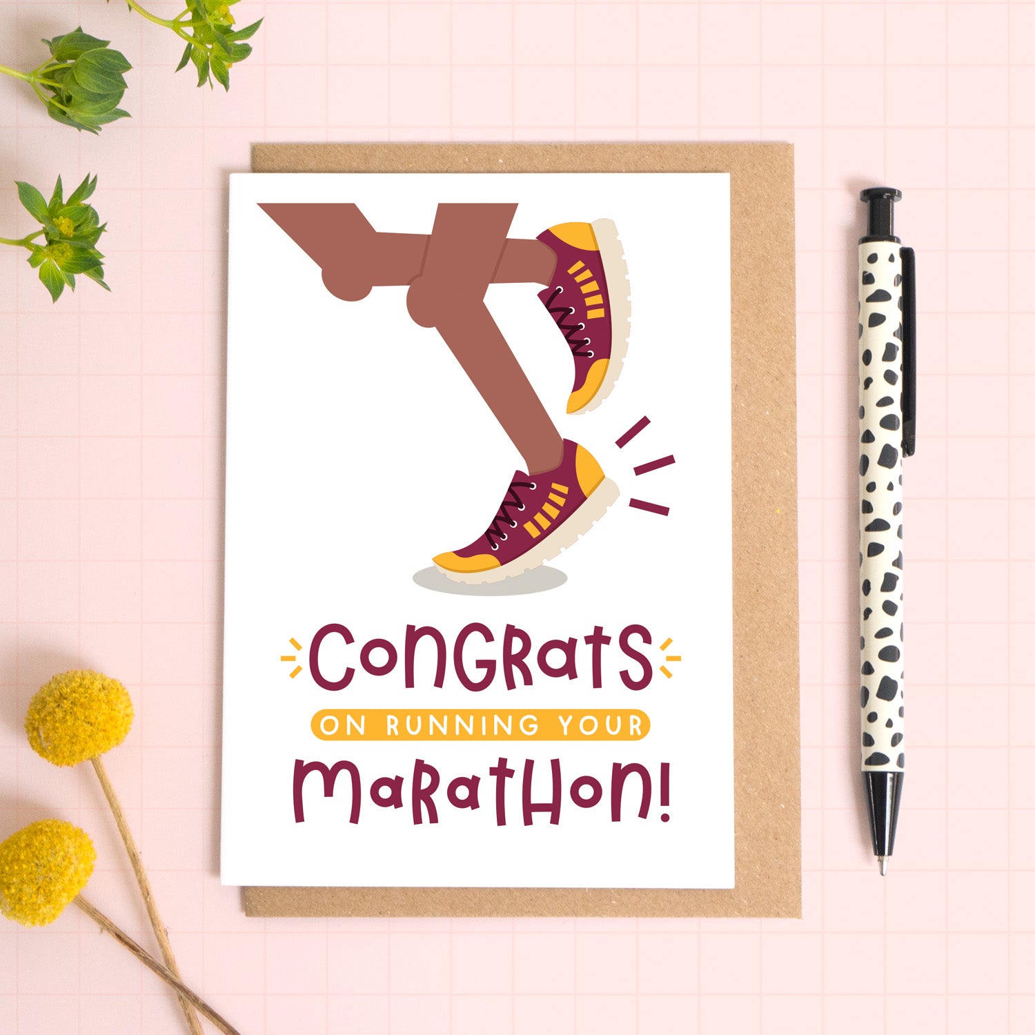 A marathon congratulations card photographed on top of a kraft brown envelope set on a pink background surrounded by foliage and a pen for scale. The card reads 'congrats on running your marathon' and features a pair of brown legs.