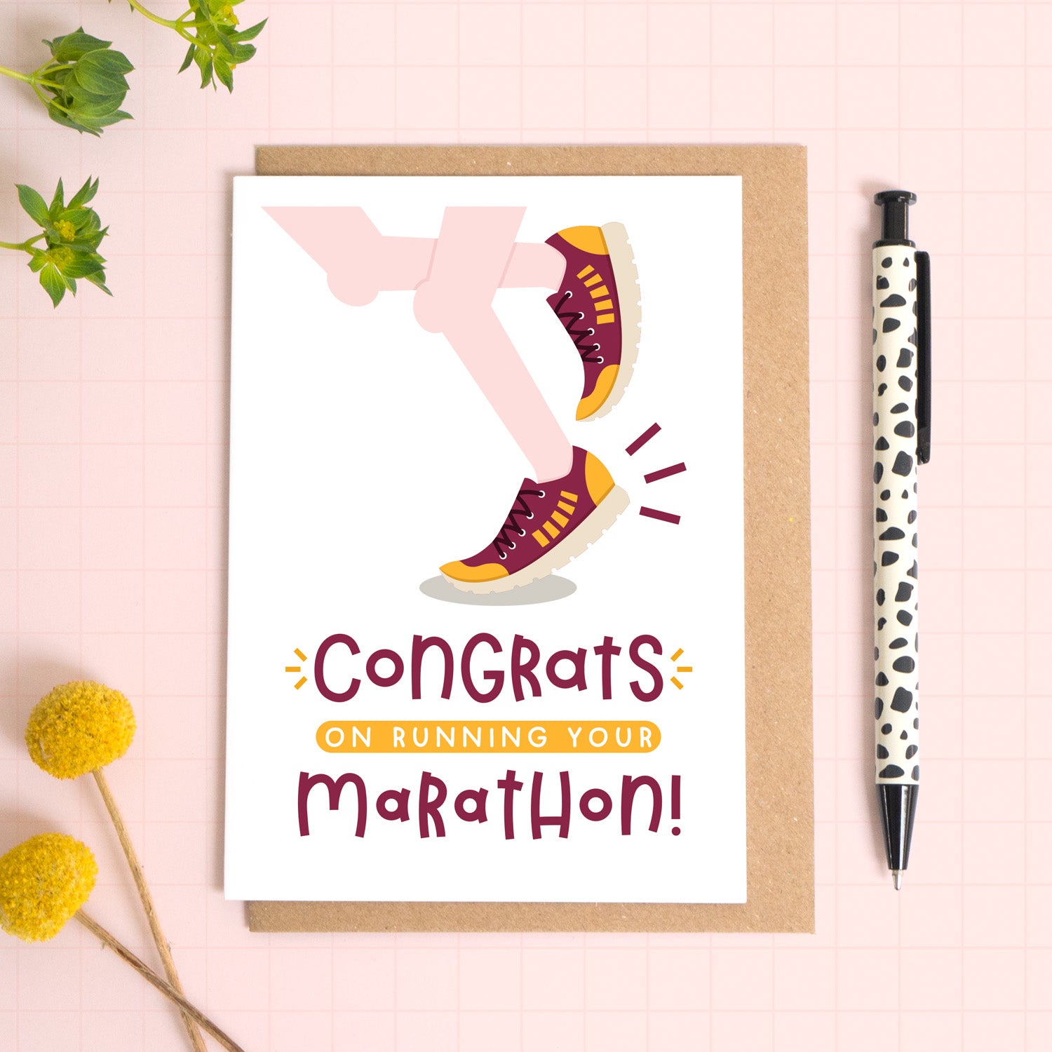 A marathon congratulations card photographed on top of a kraft brown envelope set on a pink background surrounded by foliage and a pen for scale. The card reads 'congrats on running your marathon' and features a pair of pale pink legs.