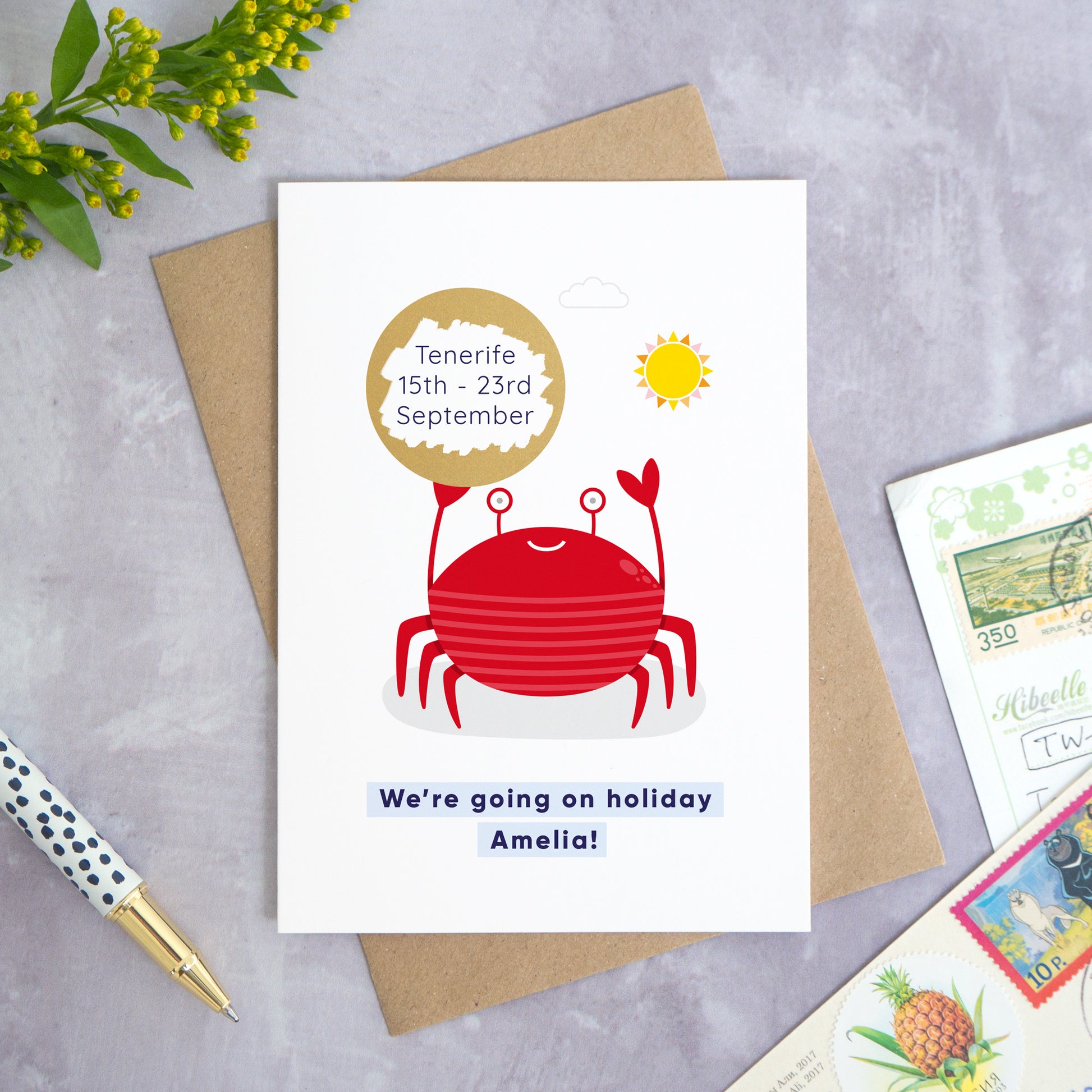 A seaside holiday reveal scratch card featuring a friendly red crab holding the scratch panel. The gold panel has been scratched off to reveal the holiday destination. Beneath the crab illustration the text reads: “we’re going on holiday Amelia”. This card has been shot on a grey background with a pen and foliage.