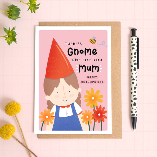 A garden gnome card with a white border featuring the phrase 'There's gnome one like you mum, happy mother's day!'. The card is laid on a kraft brown envelope on top of a pink background next to a pen and flowers.