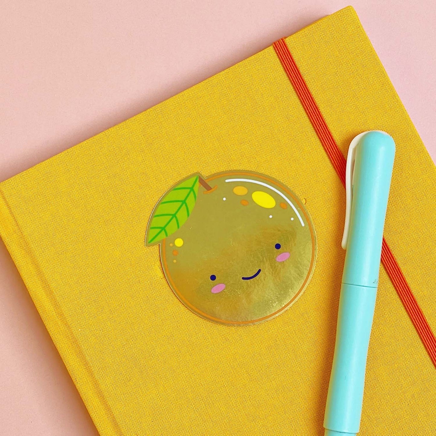 A shiny mirror effect gold sticker featuring a smiling orange lying on top of a yellow journal on a pink background