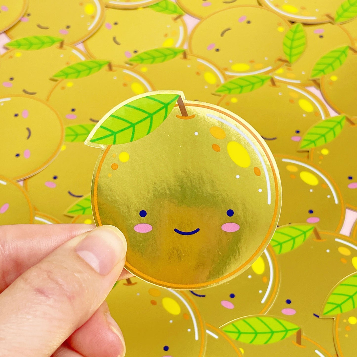 A shiny mirror effect gold sticker featuring a smiling orange being held over a pile of stickers of the same design