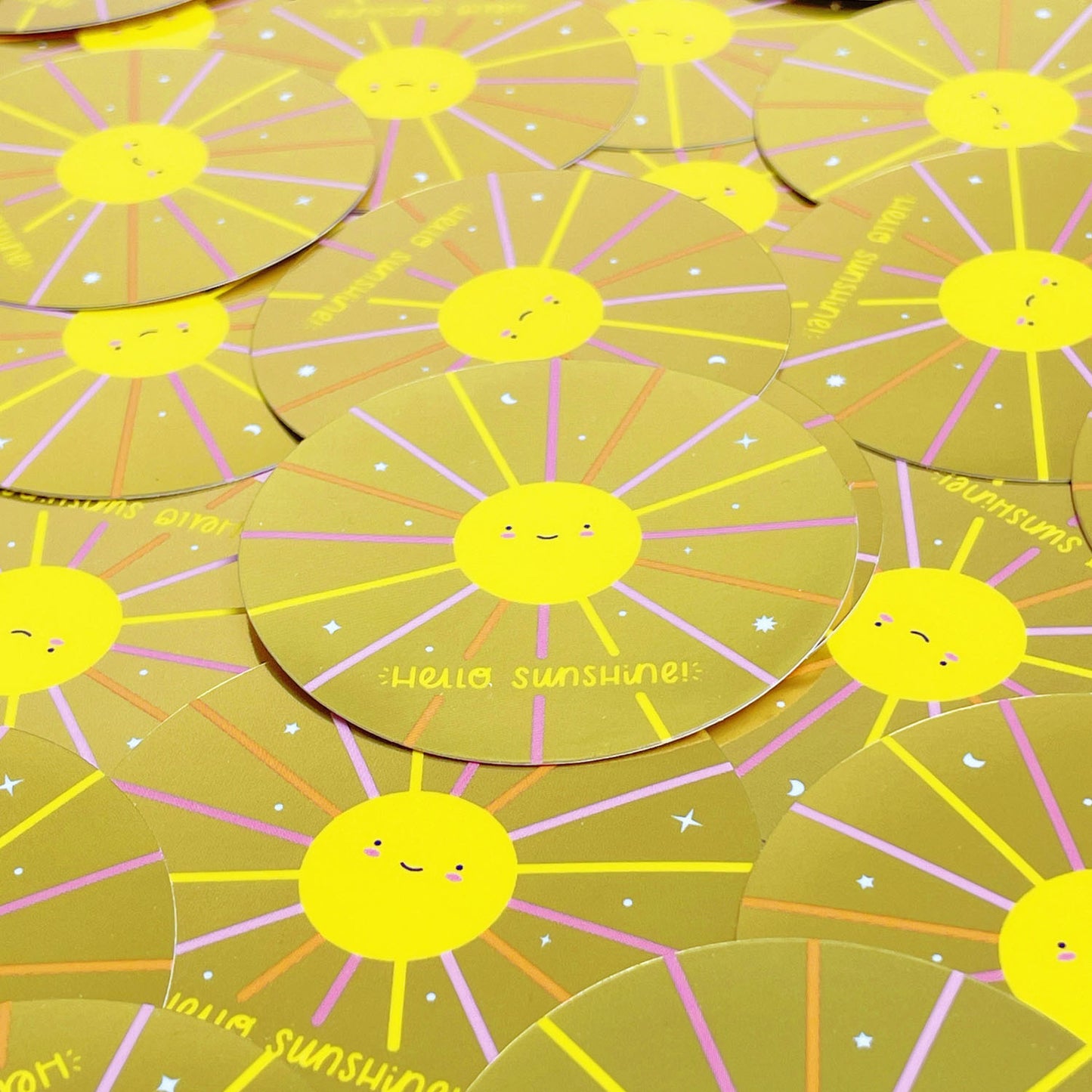 A pile of shiny mirror effect gold stickers featuring a smiling sun. The surface is covered with dozens of the same design.