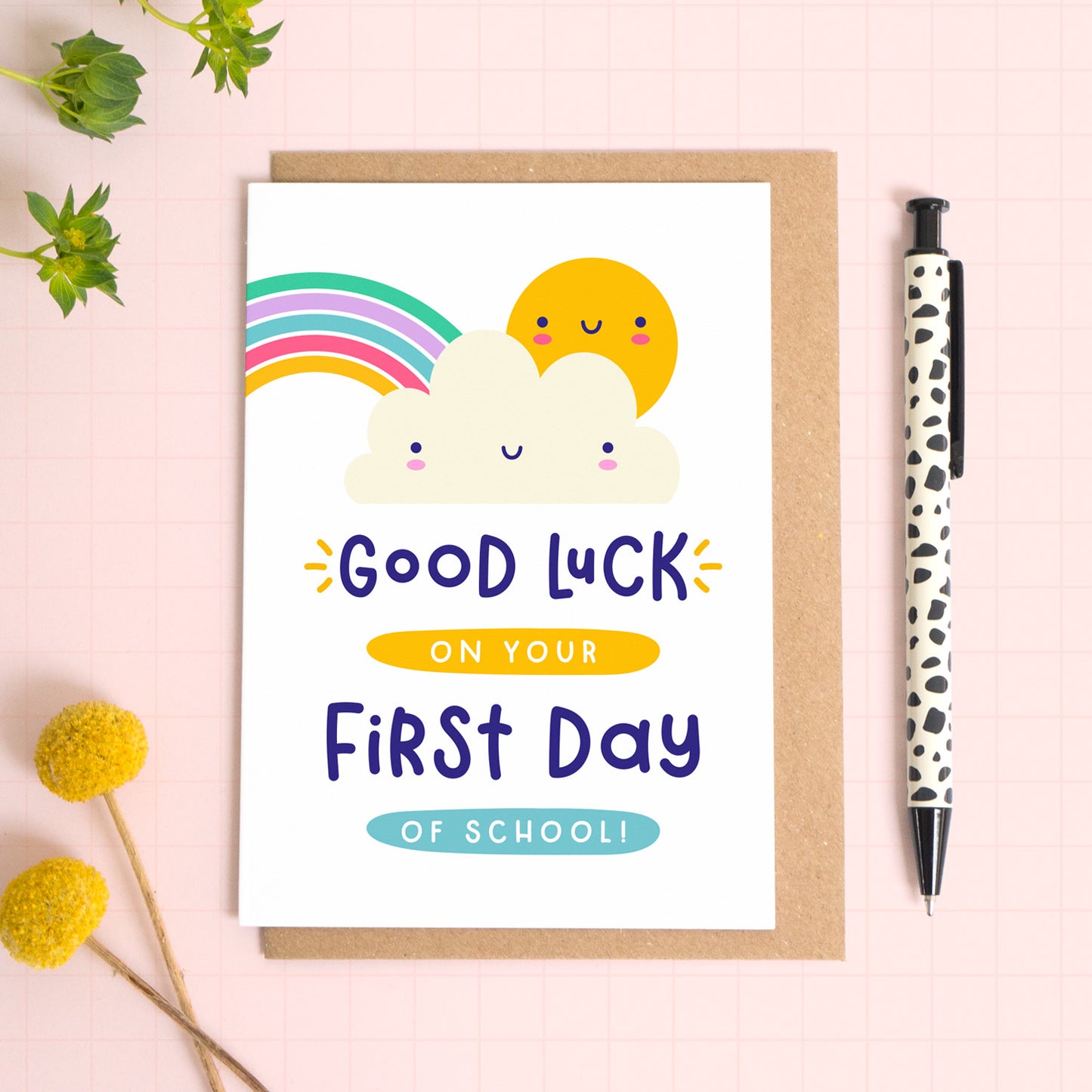 A good luck on your first day back to school card featuring a rainbow, cute cloud and happy sun. The card has been photographed on it's kraft brown envelope on a pink surface with foliage and a pen for scale.