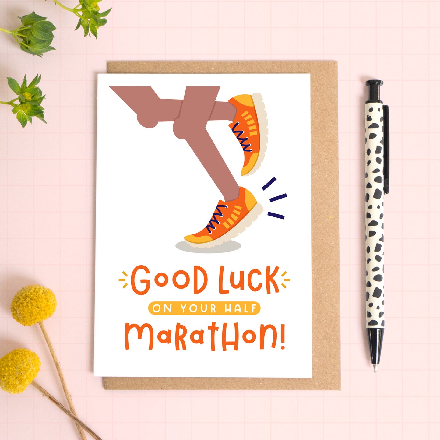 A half marathon good luck card photographed on top of a kraft brown envelope set on a pink background surrounded by foliage and a pen for scale. The card reads 'good luck on your half marathon' and features a pair of brown legs.