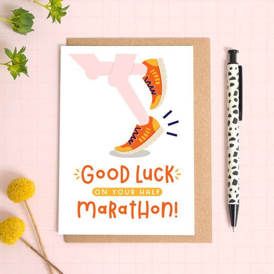 A half marathon good luck card photographed on top of a kraft brown envelope set on a pink background surrounded by foliage and a pen for scale. The card reads 'good luck on your half marathon' and features a pair of pale pink legs.