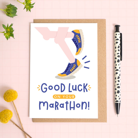 A marathon good luck card photographed on top of a kraft brown envelope set on a pink background surrounded by foliage and a pen for scale. The card reads 'good luck on your marathon' and features a pair of pale pink legs.