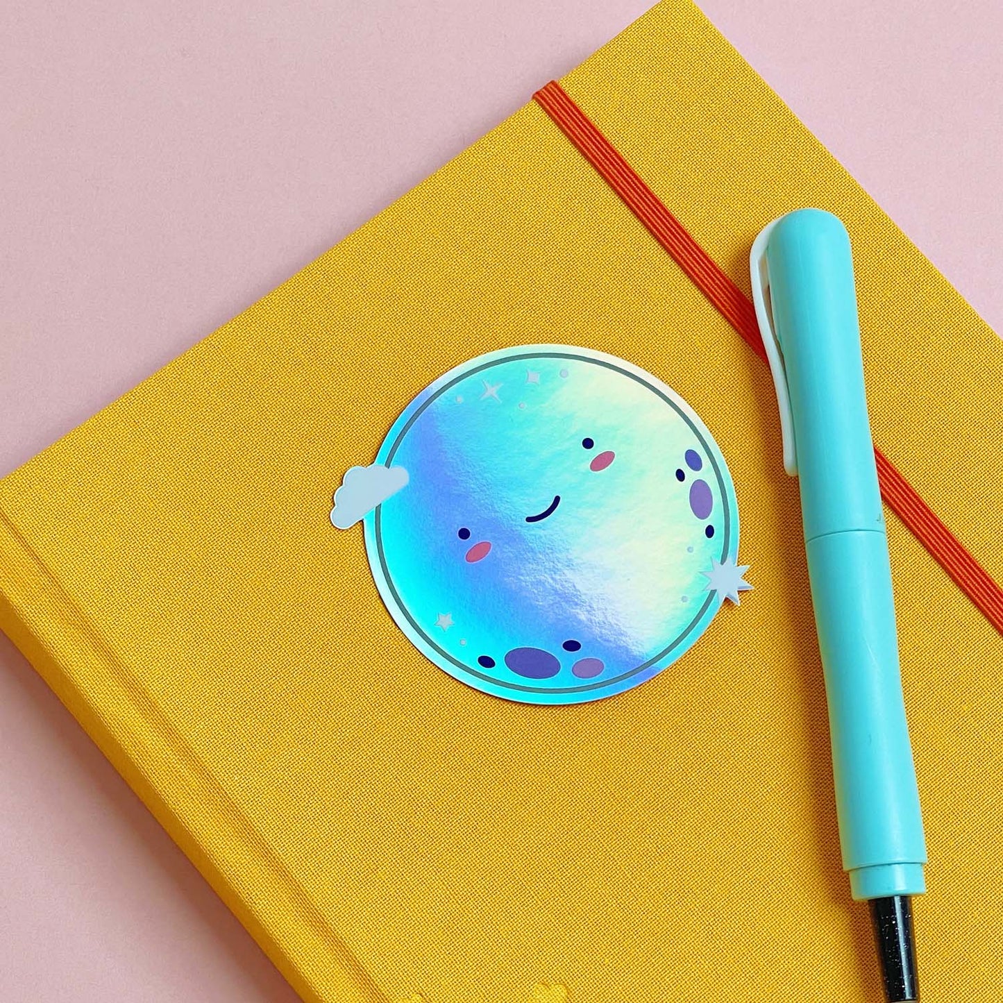 A shiny iridescent holographic vinyl sticker featuring a smiling moon lying on top of a yellow journal on a pink background