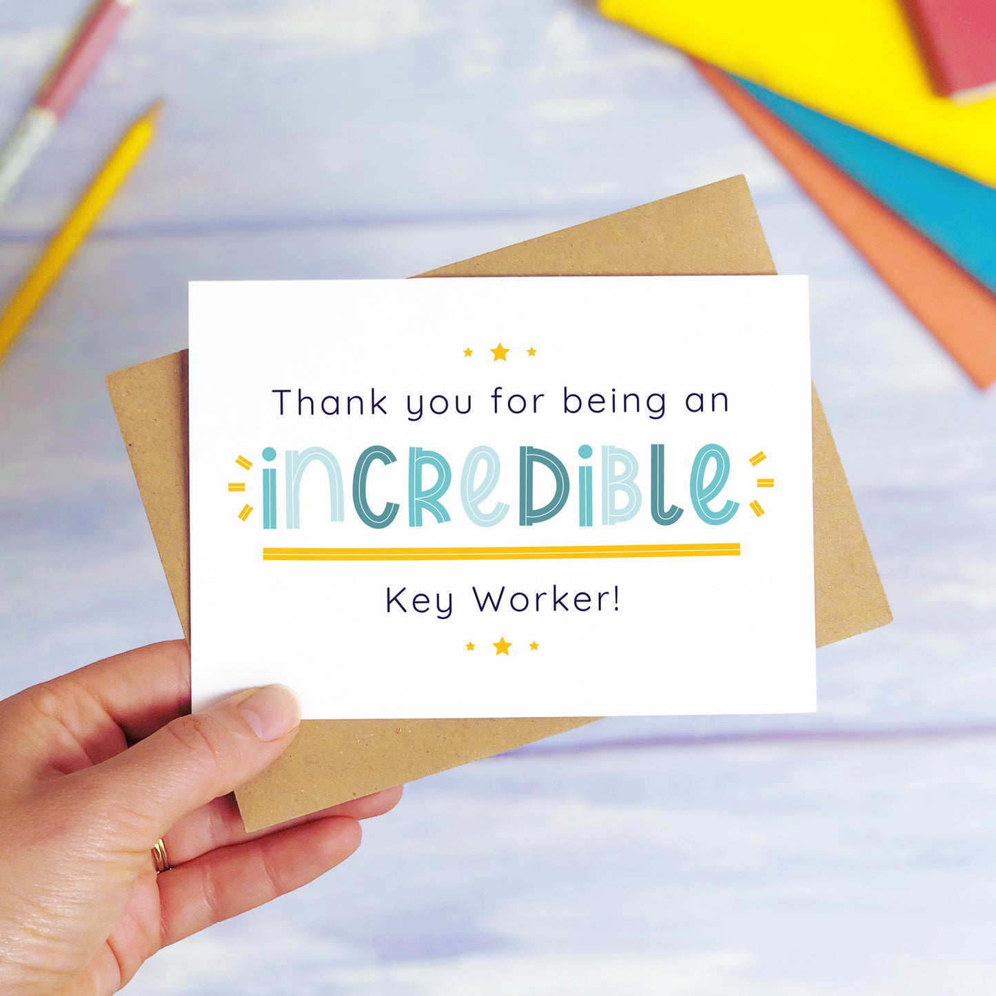 An incredible key worker nursery thank you card being held over a blue background with colourful text books and two pens. The card is sat on top of its kraft brown envelope. This is the teacher card with the blue text colour option.