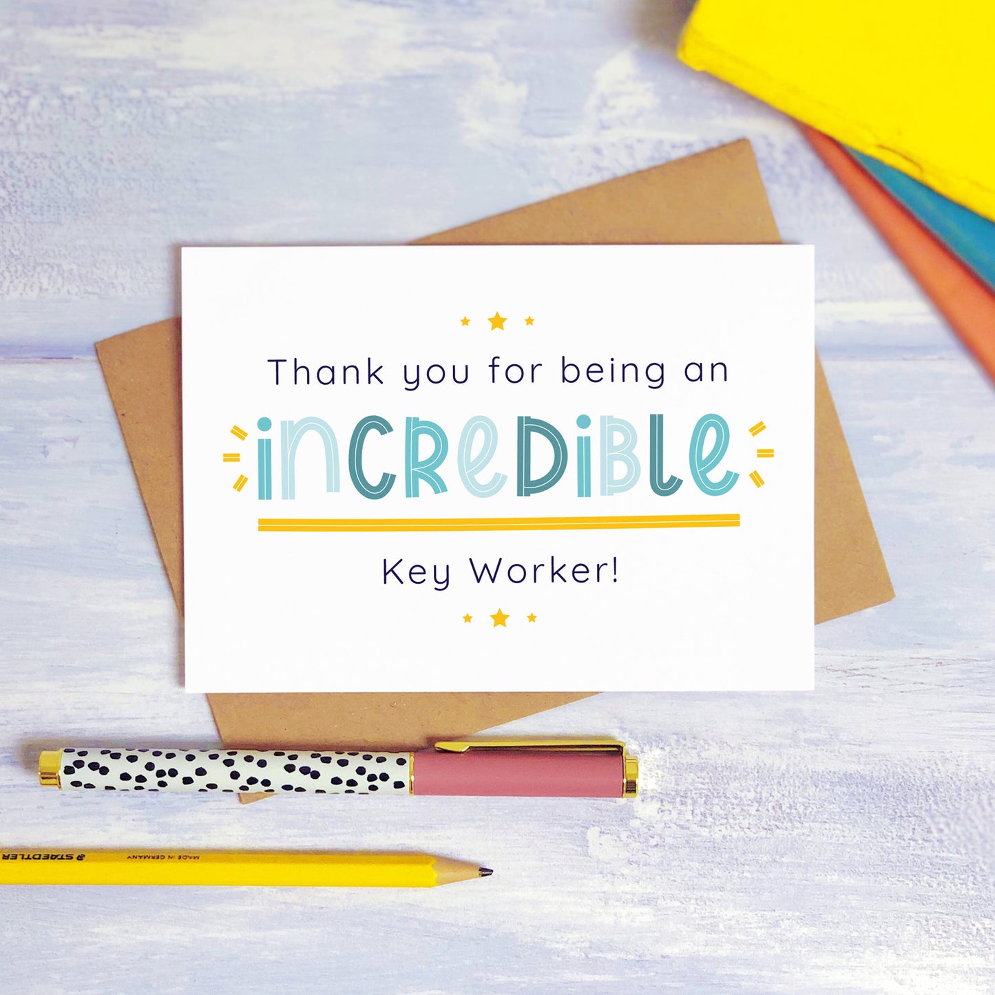 An incredible key worker nursery thank you card lying on top of a kraft brown envelope, flat lay style on a blue textured background with colourful text books, a pen and a pencil. This teacher card features the blue text colour option.