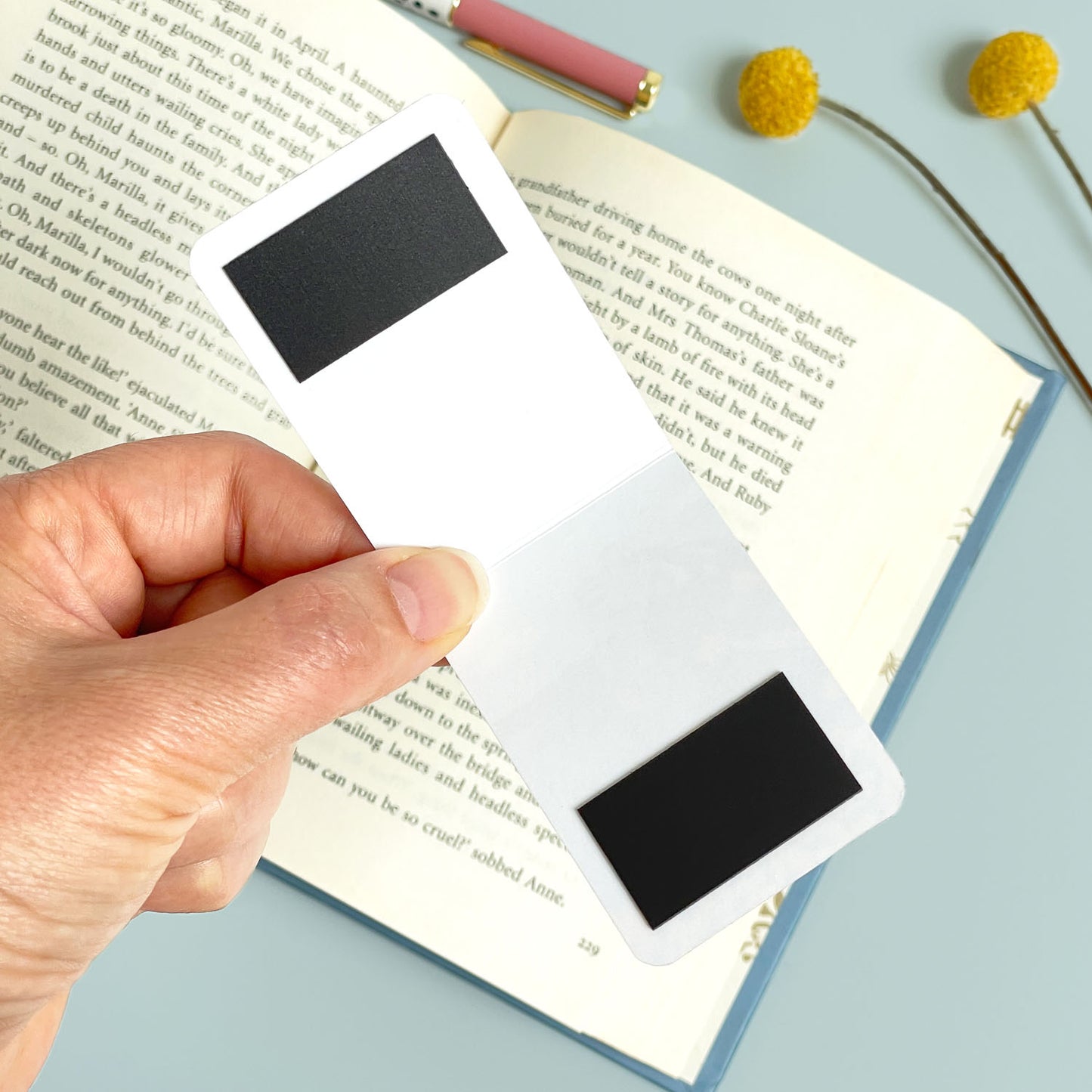 Hand holding open the magnetic bookmark over an open book with a pen and flowers on a light blue background.