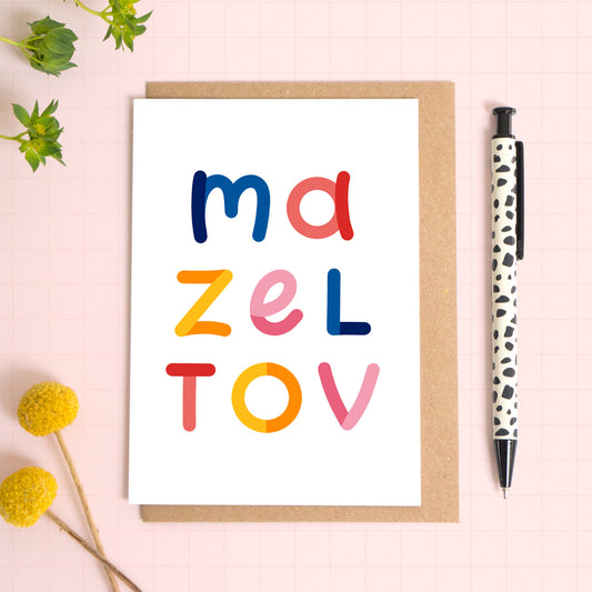 This Mazel Tov card has been photographed on top of a kraft brown envelope sitting upon a pink background with foliage and flowers to the left and a spotty pen to the right.