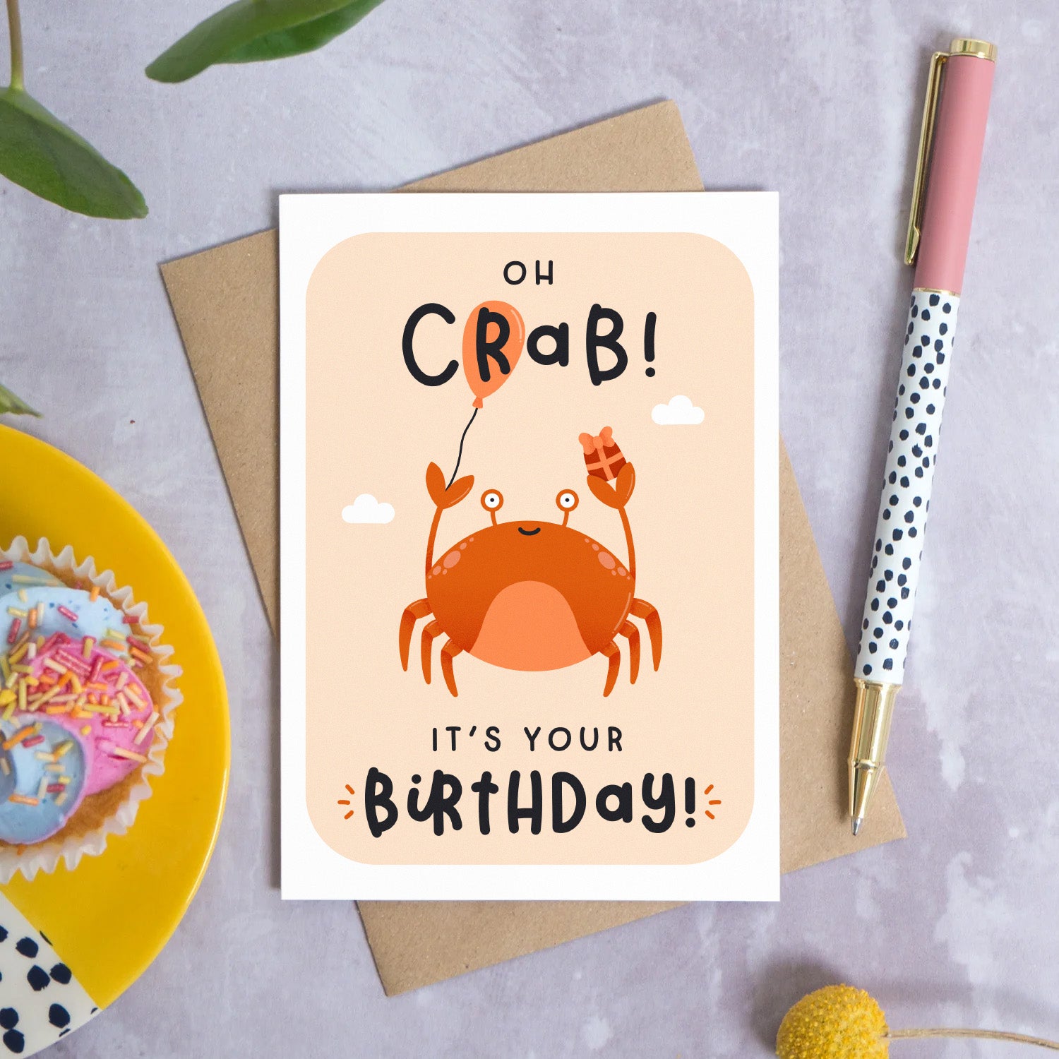 A birthday card featuring a crab holding a balloon and a present with the words ‘oh crab, it’s your birthday!’. The card is lying on a Kraft brown envelope on a grey surface. Surrounding the card are leaves, flowers, a pen and a cupcake on a plate.