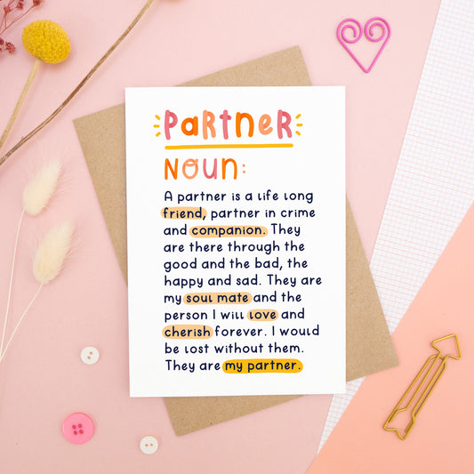 A heartfelt, colourful dictionary definition style card with the definition of a partner, photographed on a pink background with floral props, paper clips, and buttons. 