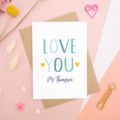 A personalised love you card photographed on a pink background with floral props, paper clips, and buttons. This image shows the ‘love you’ anniversary card in the blue colour way. The personalisation is navy and there are pops of yellow in the hearts.