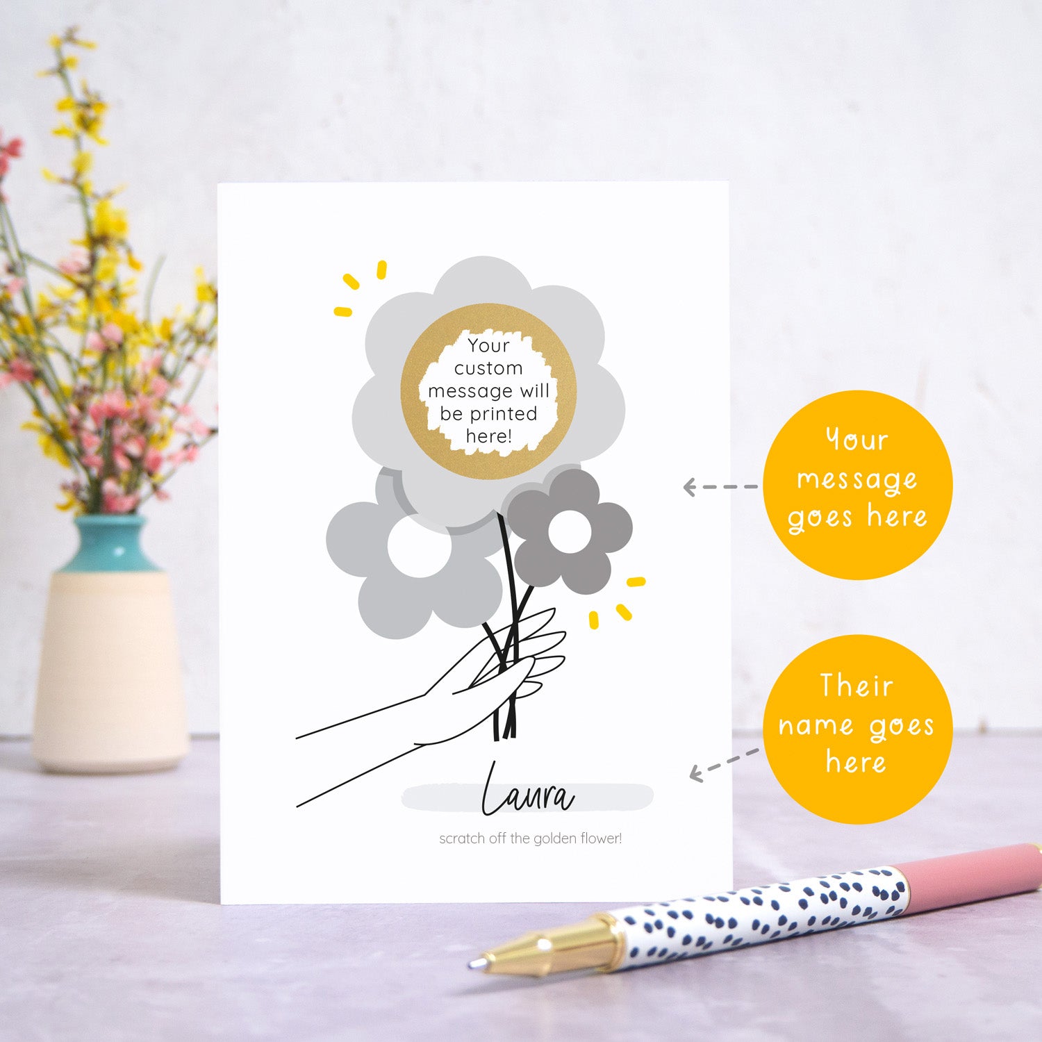 The scratch and reveal card has been photographed standing up on top of a grey surface. There is a small jar of flowers in the background and a pen in the foreground. To the right of the card are two circles which point to the areas that can be personalised. These are the name and the scratch off message. The gold circle has been scratched off to reveal the custom text.