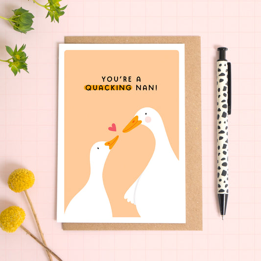 A peach card with a white border featuring two cute ducks and the phrase 'you're a quacking nan!'. The card is laid on a kraft brown envelope on top of a pink background next to a pen and flowers.