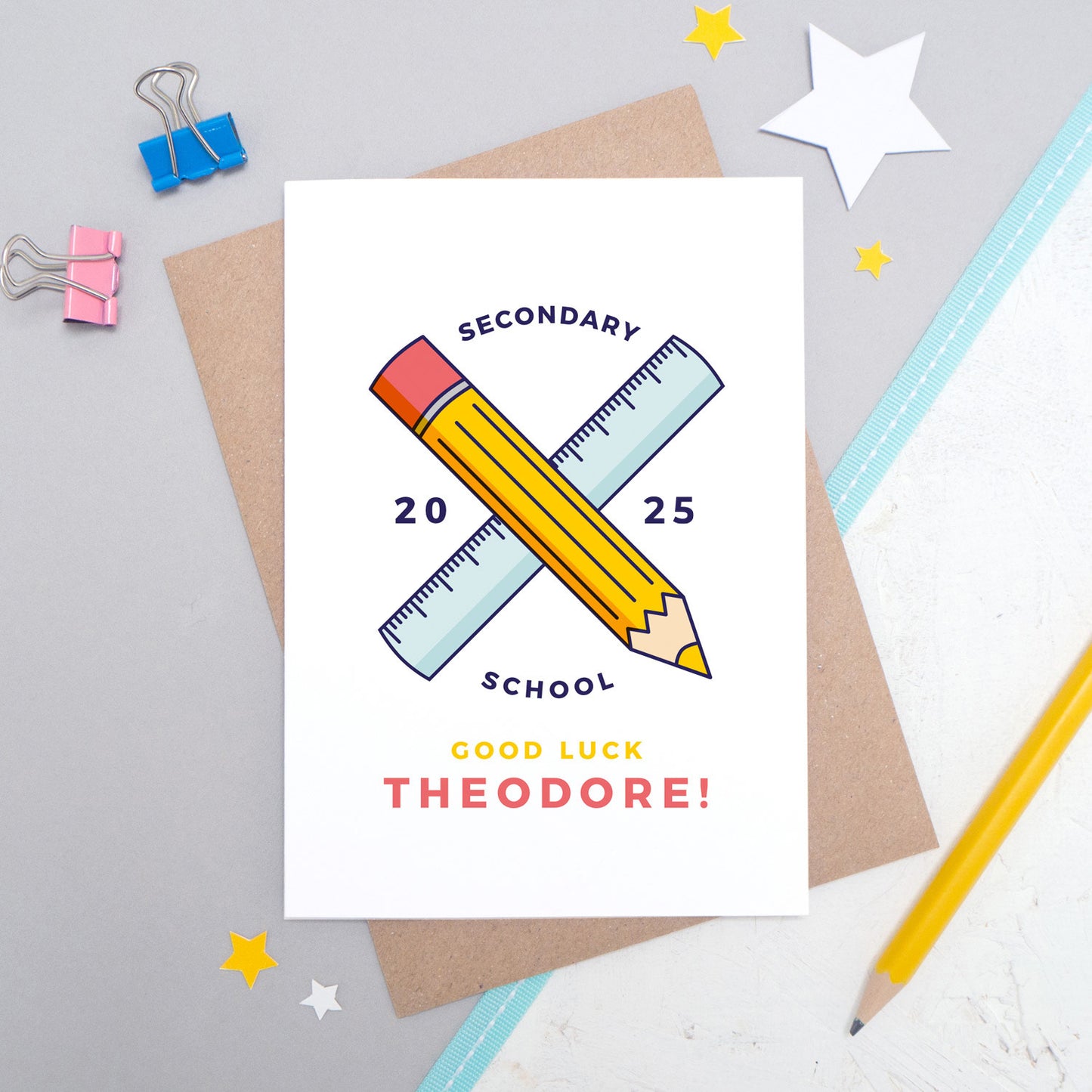 A good luck for secondary, personalised back to school card personalised with a young persons name, and a pencil and ruler illustration. The card is laid on it's kraft brown envelope on a grey and white background.