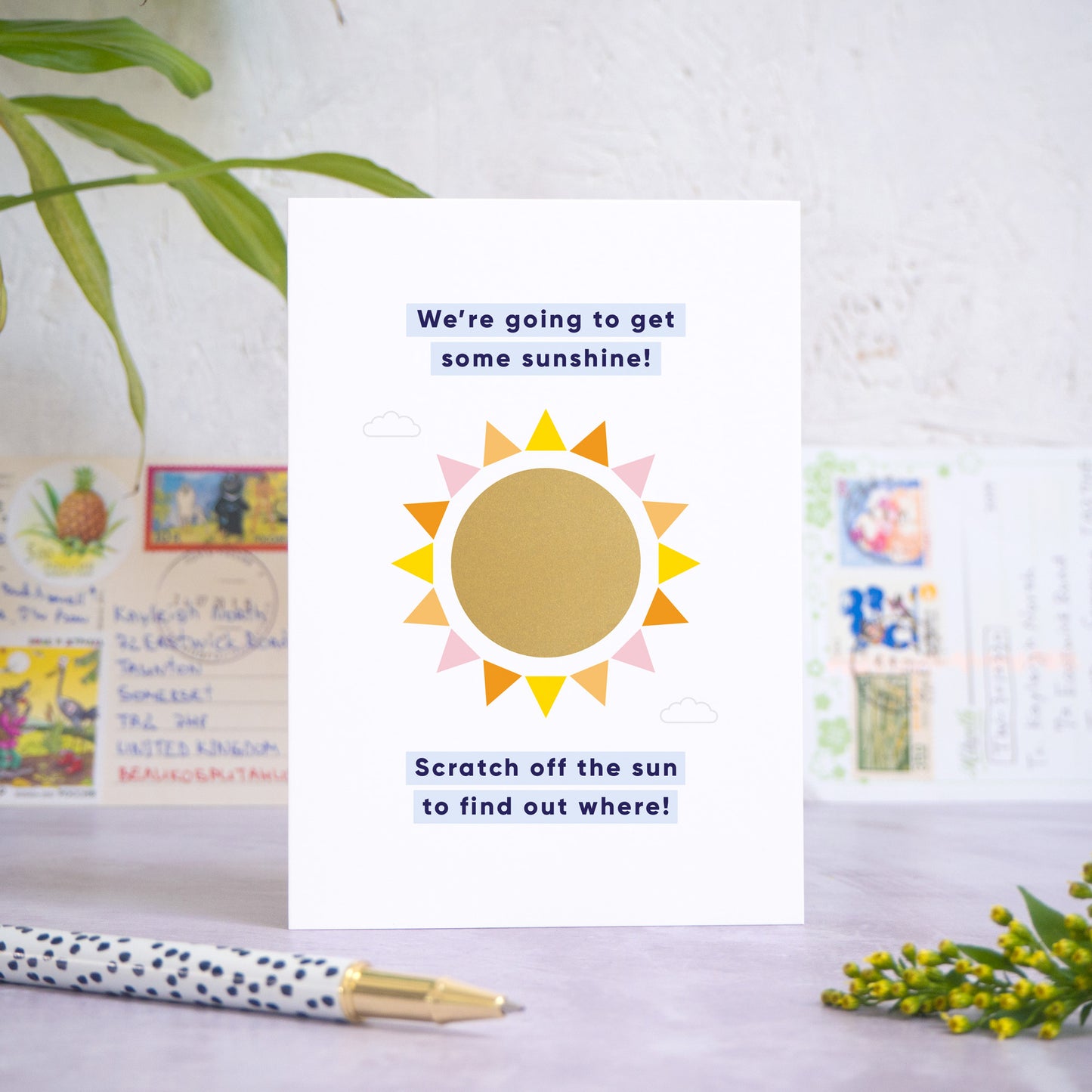 A holiday reveal scratch card featuring a shiny gold sun. The gold panel has not yet been scratched off to reveal the holiday destination. The text reads: “We're going to get some sunshine! Scratch off the sun to find out where!”. The card is standing on a grey surface with postcards in the background and a pen and foliage in the foreground.