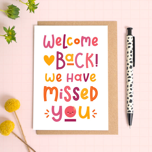 This joyful welcome back card has been photographed on top of a kraft brown envelope sitting upon a pink background with foliage and flowers to the left and a spotty pen to the right.