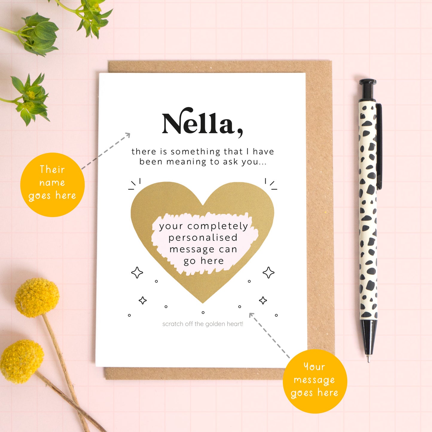 A ‘will you be my birthing partner’ scratch and reveal card photographed lying on a Kraft brown envelope, set upon a pink background surrounded by foliage and a pen for scale. The gold panel has been scratched off to reveal the personalised question and there are orange circles pointing to the areas that can be personalised. These include the name and the scratch off message.