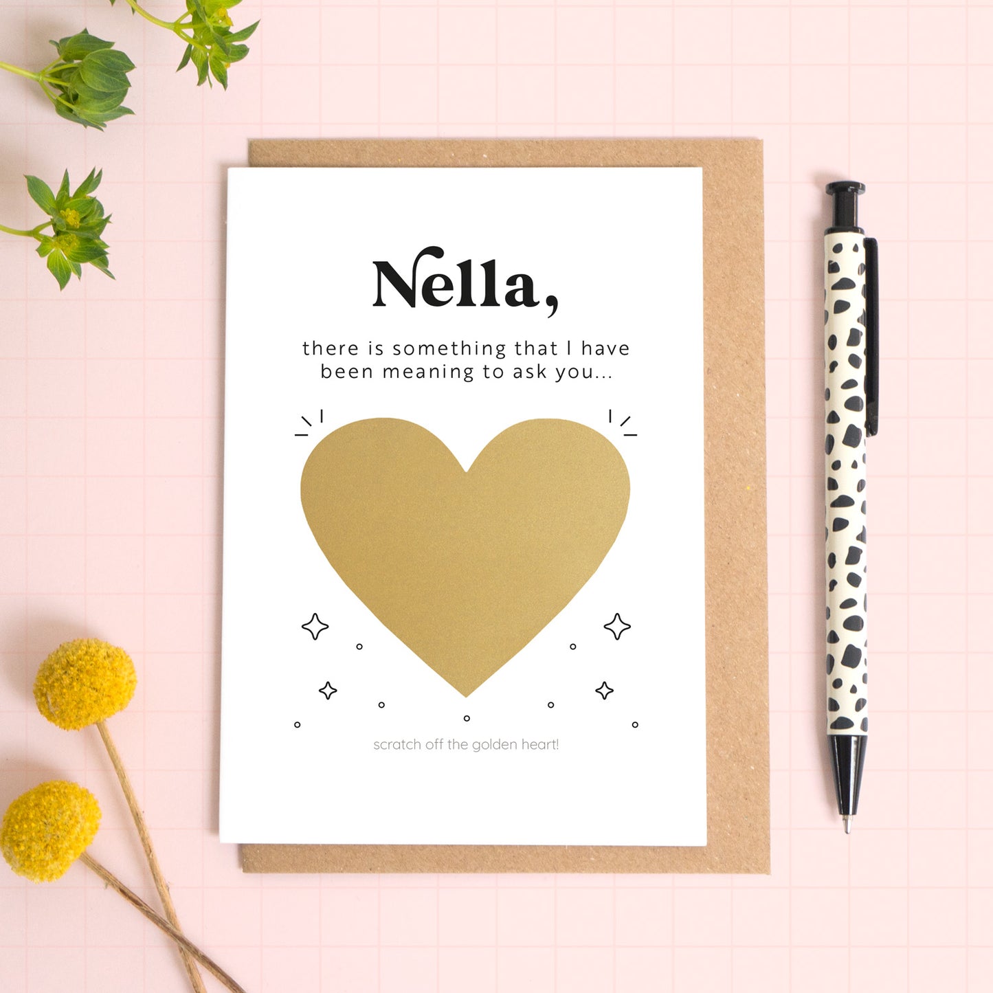 A ‘will you be my birthing partner’ scratch and reveal card photographed lying on a Kraft brown envelope, set upon a pink background surrounded by foliage and a pen for scale. The gold panel has not yet been scratched off.