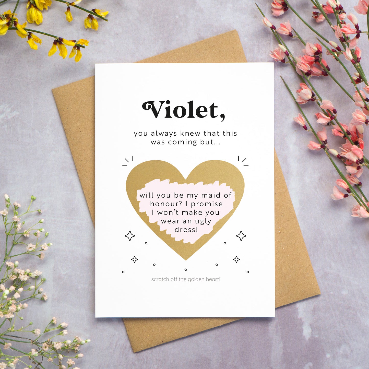 The personalised question scratch card has been photographed lying on top of a Kraft brown envelope which is on top of a grey surface surrounded by colourful flowers. The gold heart scratch panel has been scratched away to reveal the surprise question.