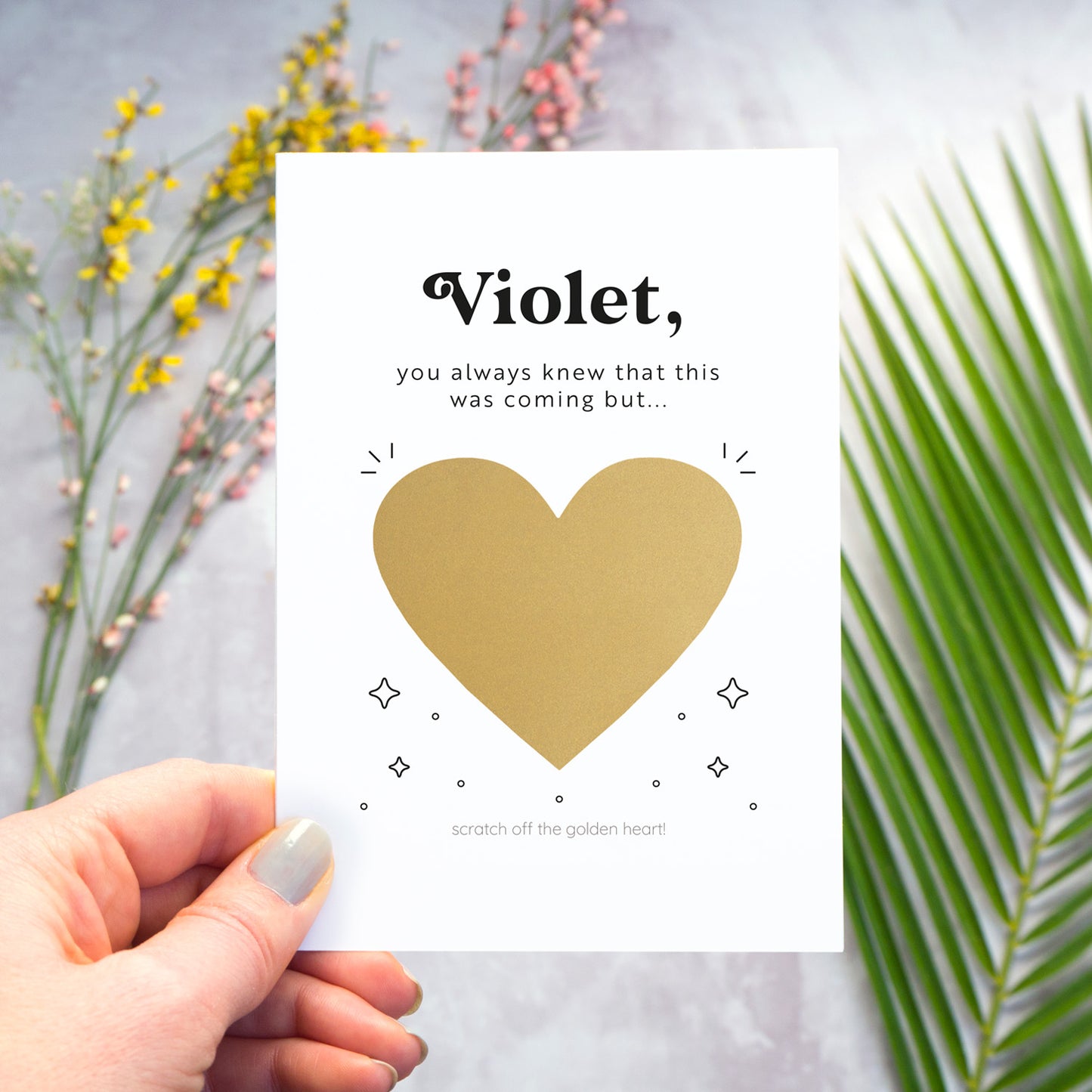 This image shows how the personalised question scratch card looks before the recipient scratches away the heart. In this image the card is being held over a grey surface which has a variety of colour flowers and foliage in the background. The heart has not yet been scratched away.
