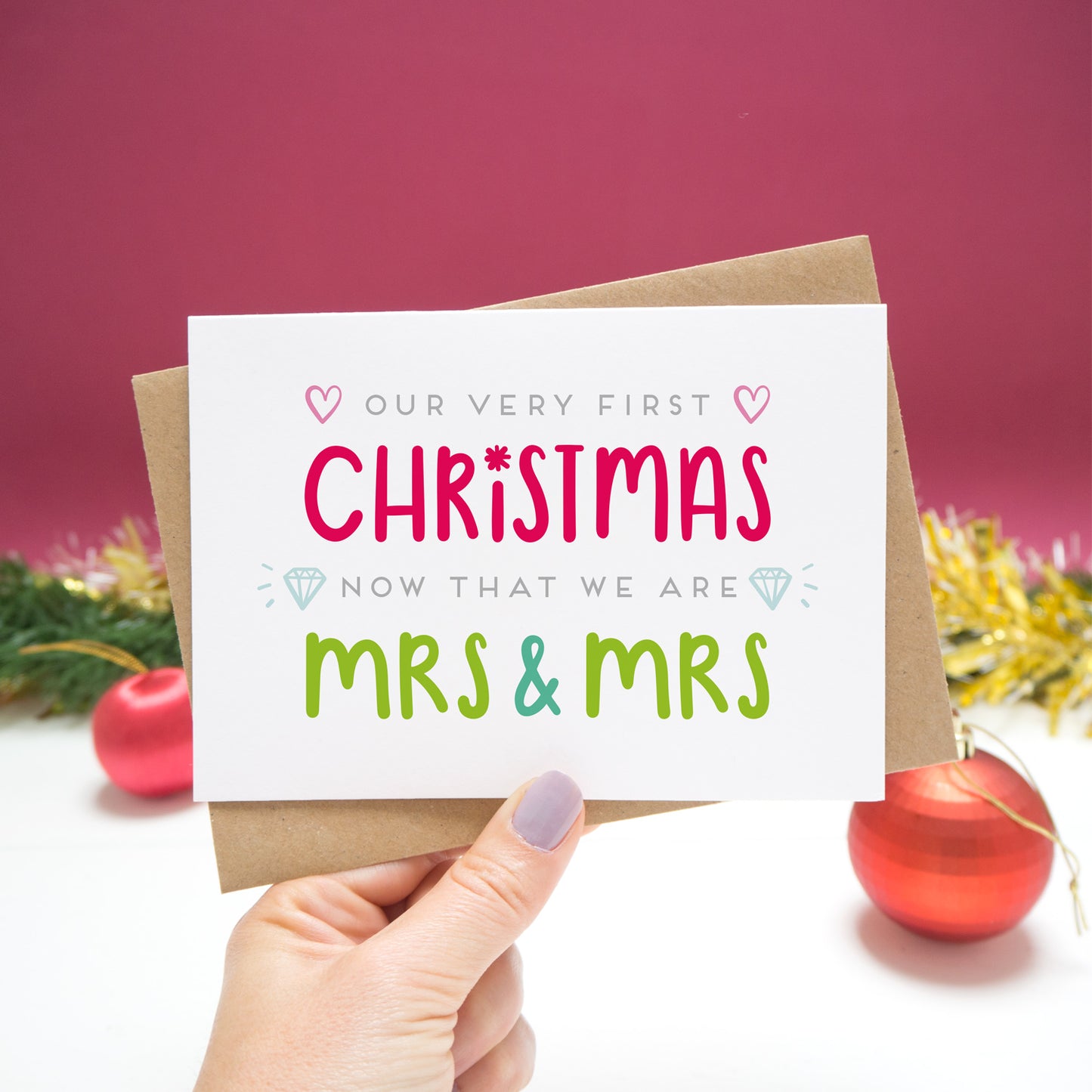 'Our very first Christmas now that we are Mrs and Mrs.' Christmas Card held in front of a Christmassy scene with baubles and tinsel.