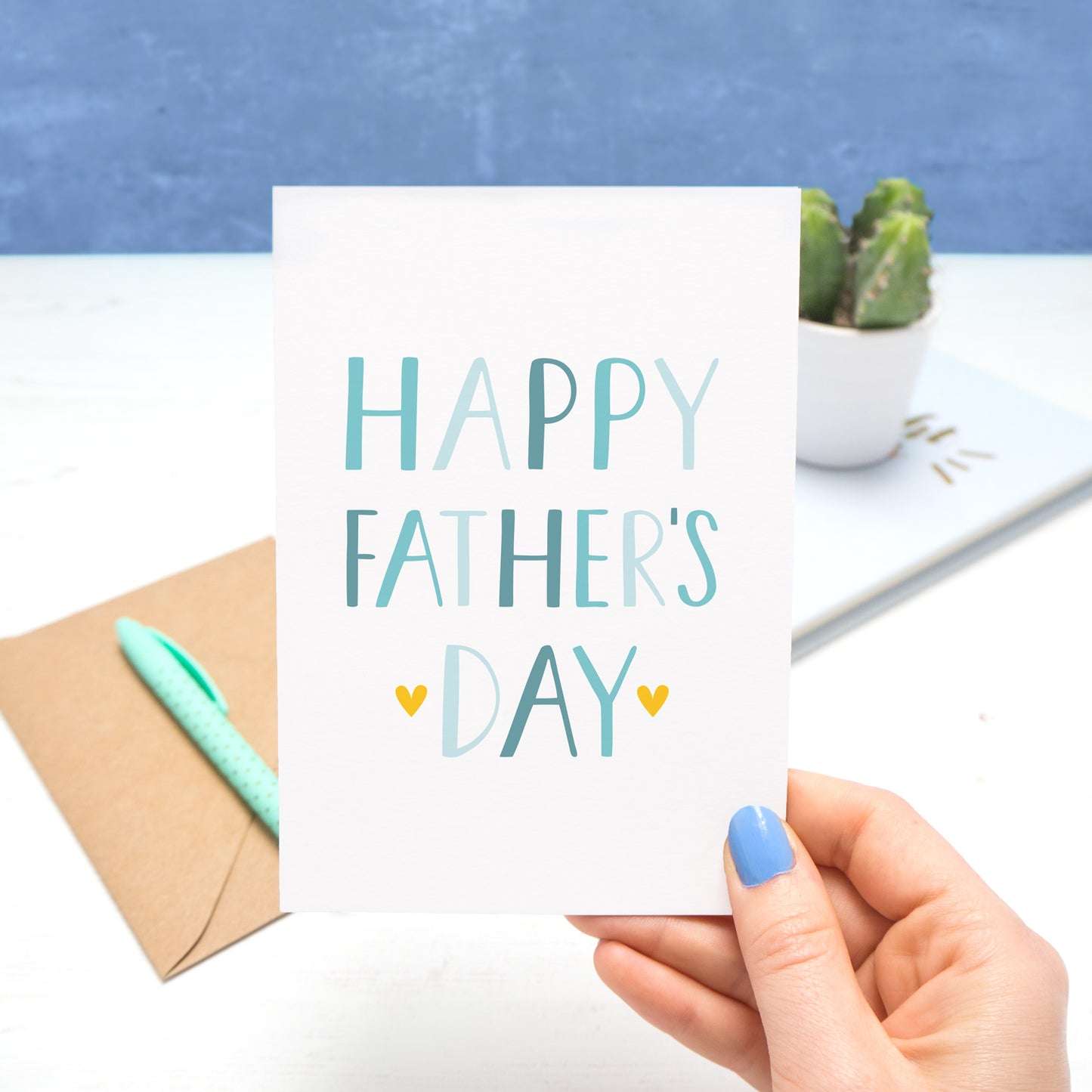 A very simple card that reads 'Happy Father's Day' held by Joanne Hawker in her Somerset Studio over a white and blue background. The text on the card is in varying shades of blue with two small yellow hearts around the word 'day'.