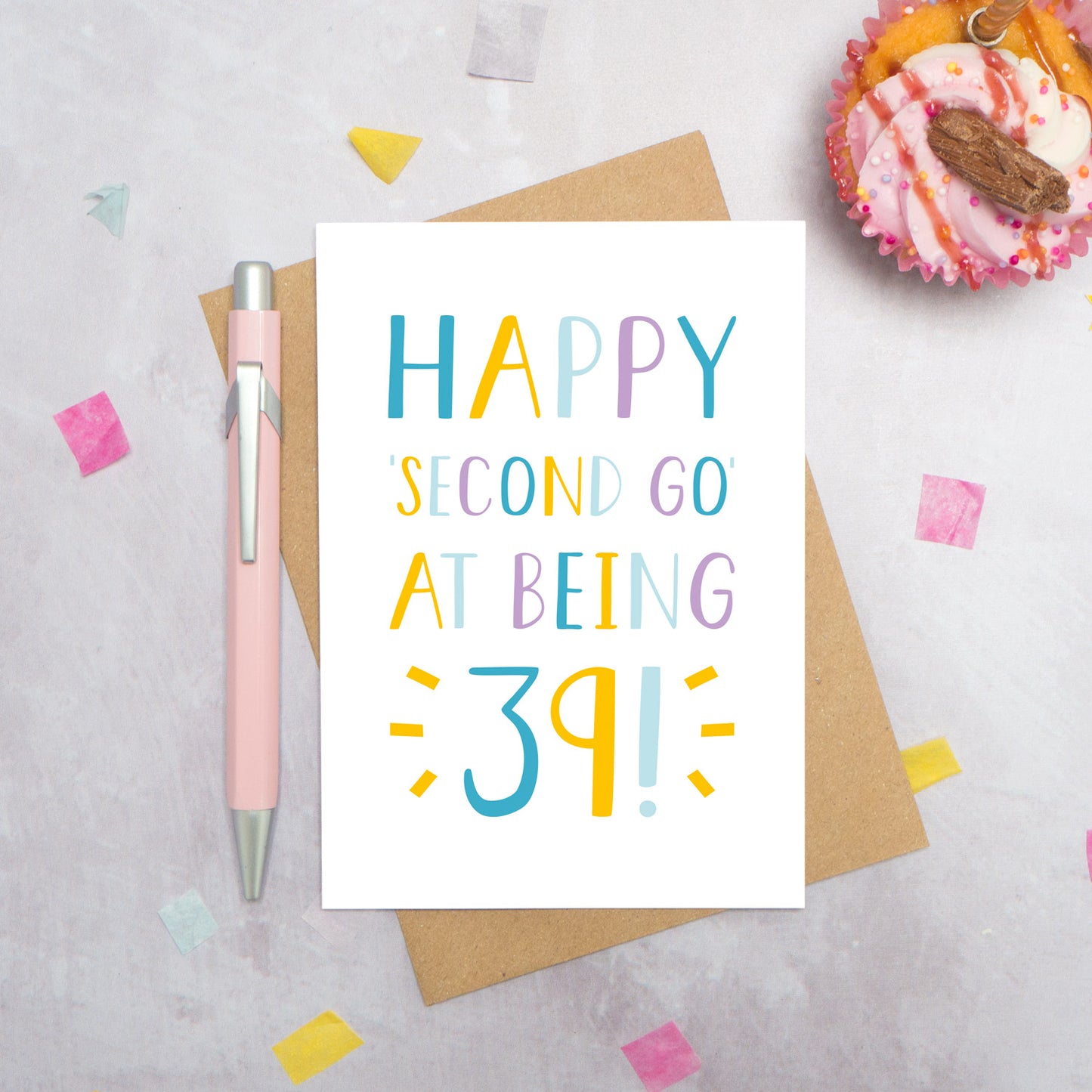 Happy second go at being 39 - milestone age card in blue, yellow and purple photographed on a grey background surrounded by a cupcake, pen and confetti.