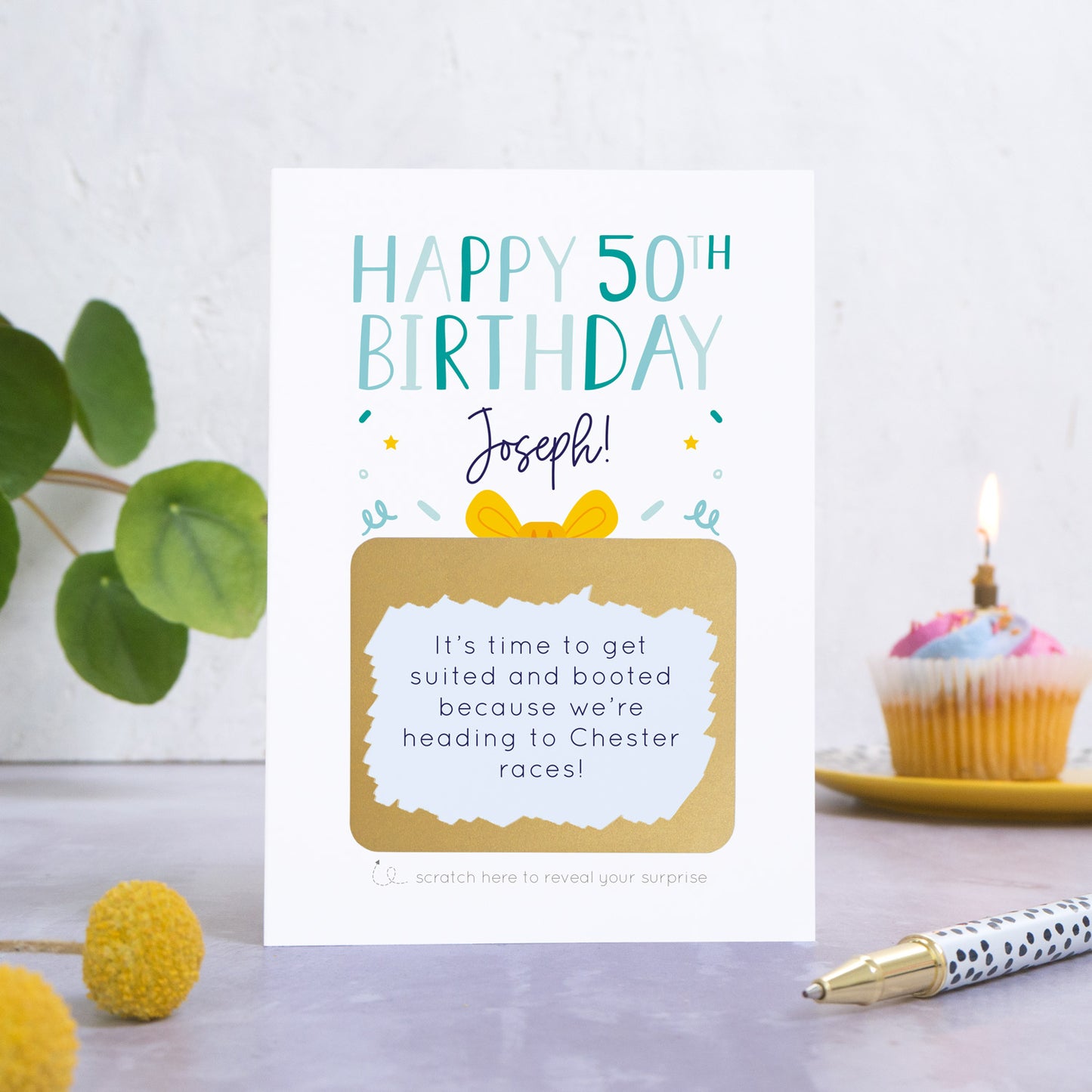 A personalised happy 50th birthday scratch card in blue that has been photographed on a white and grey background. There is foliage and yellow flowers on the left and a cupcake and a pen to the right. The card features the recipients name and a scratch panel that has been scratched off revealing a personalised message.