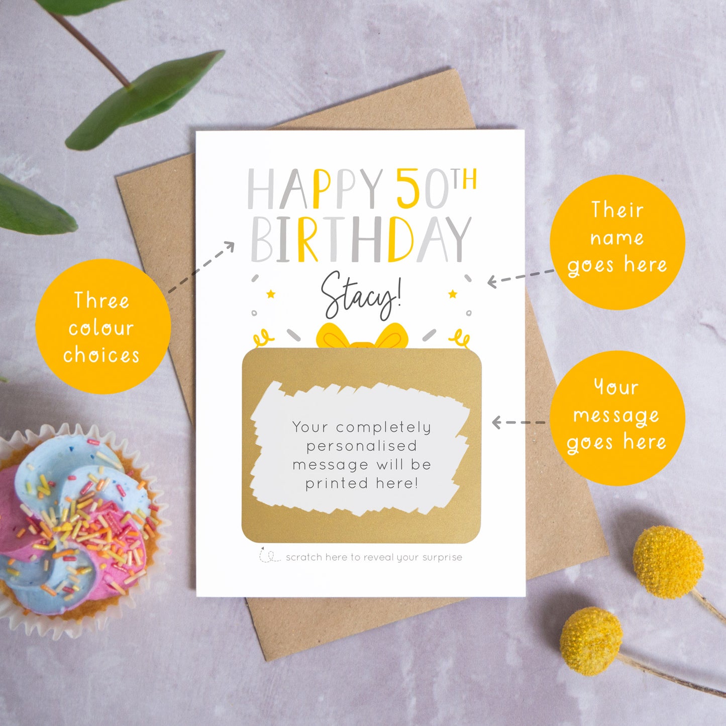 A personalised happy 50th birthday scratch card in grey that has been photographed on a grey background with foliage and a cupcake. There are also orange circles laid over the top of this image with arrows pointing to the areas that can be customised. In this instance they point to the name, the colour choices and the personalised scratch off message.