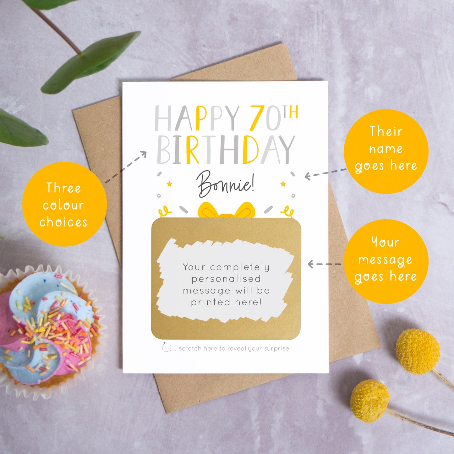 A personalised happy 70th birthday scratch card in grey that has been photographed on a grey background with foliage and a cupcake. There are also orange circles laid over the top of this image with arrows pointing to the areas that can be customised. In this instance they point to the name, the colour choices and the personalised scratch off message.