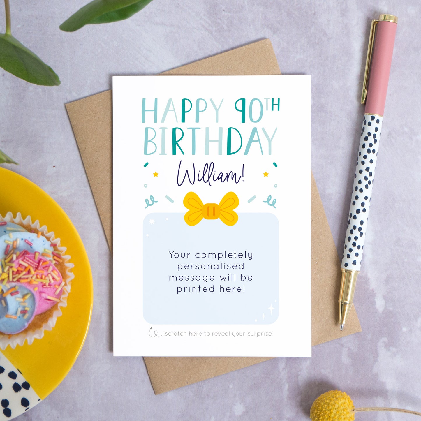 A personalised happy 90th birthday scratch card that has been photographed flat lay style on a grey concrete style background surrounded with foliage, a cupcake and a pen. The card itself shows how it would look once the gold panel has been completely removed.