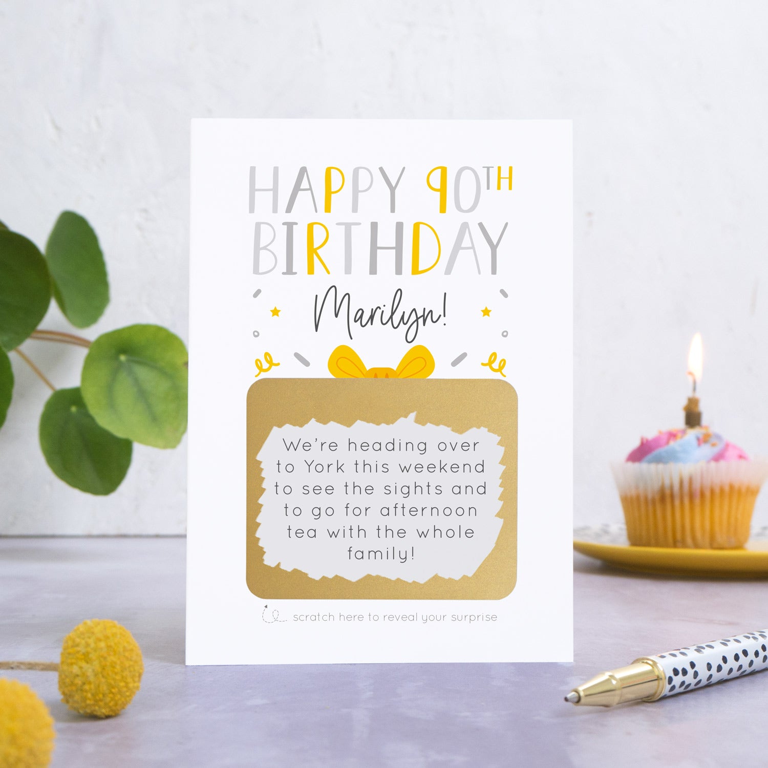 A personalised happy 90th birthday scratch card in grey that has been photographed on a white and grey background. There is foliage and yellow flowers on the left and a cupcake and a pen to the right. The card features the recipients name and a scratch panel that has been scratched off revealing a personalised message.