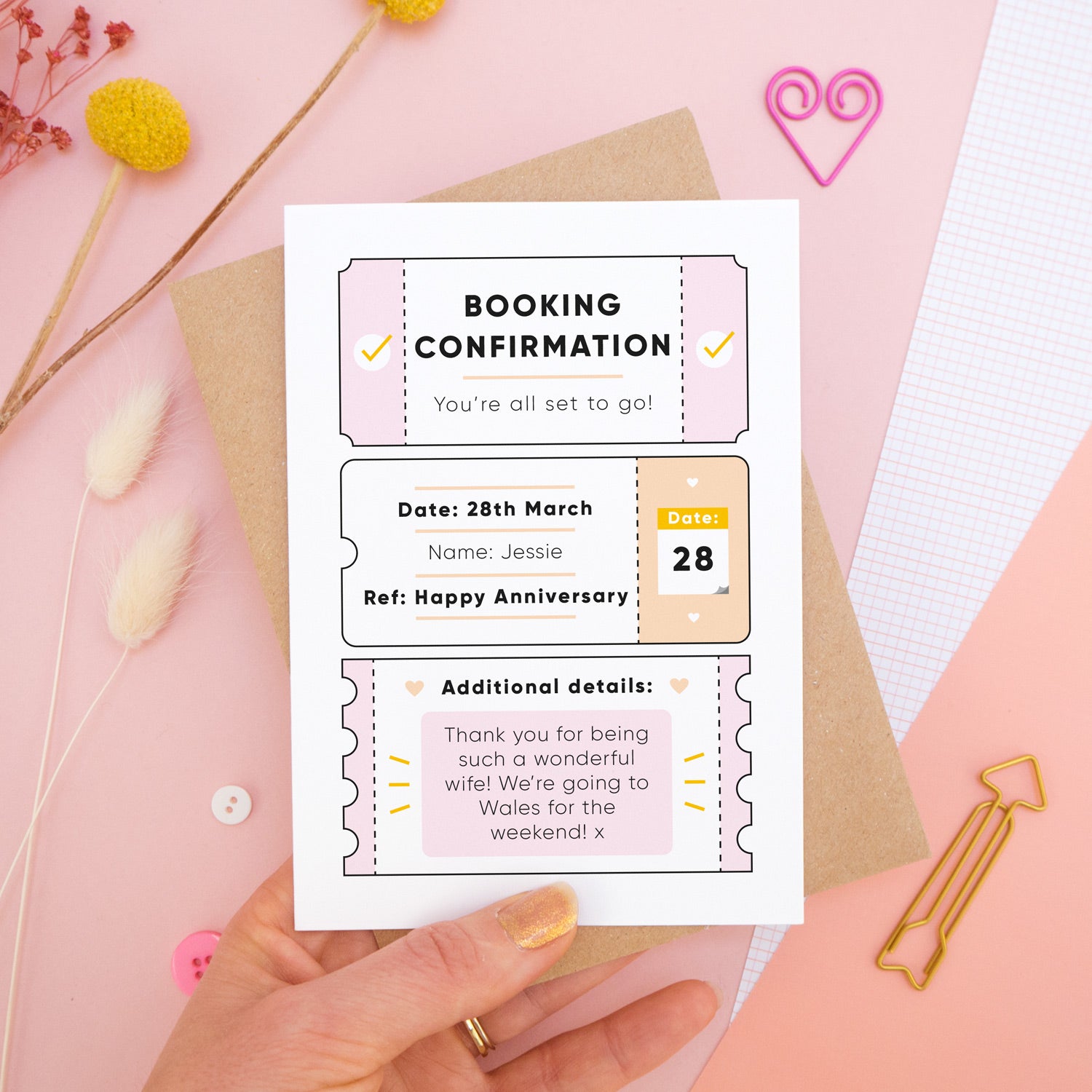A personalised anniversary booking confirmation gift card held in front of a pink background scattered with dry flowers, grid paper, buttons and a heart. Pictured is the pink version of the card on top of a kraft brown envelope.