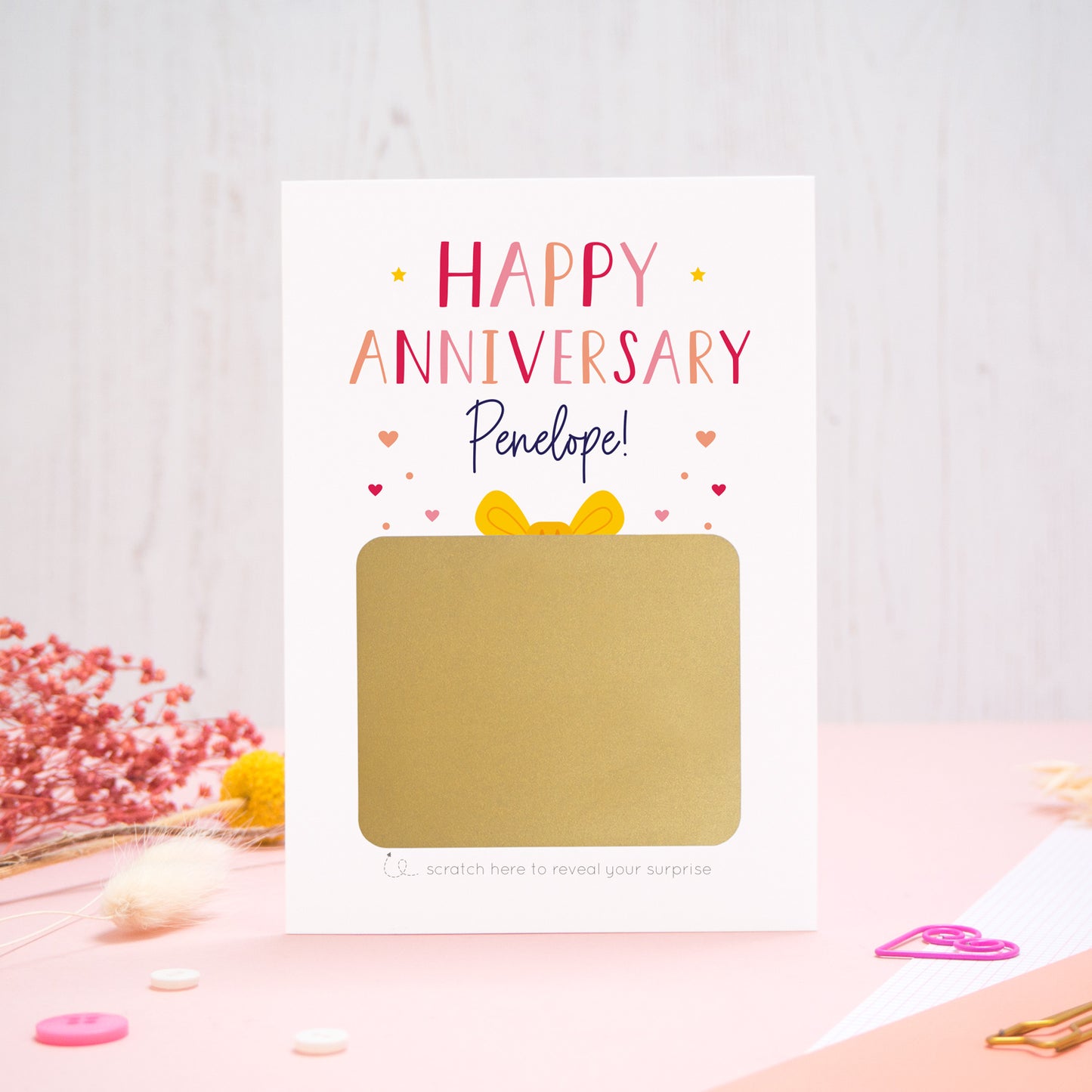 A personalised happy anniversary scratch card photographed on a pink and white background with floral props, paper clips, and buttons. The card has not yet been scratched off to reveal the hidden message and is in the red & peach colour scheme.