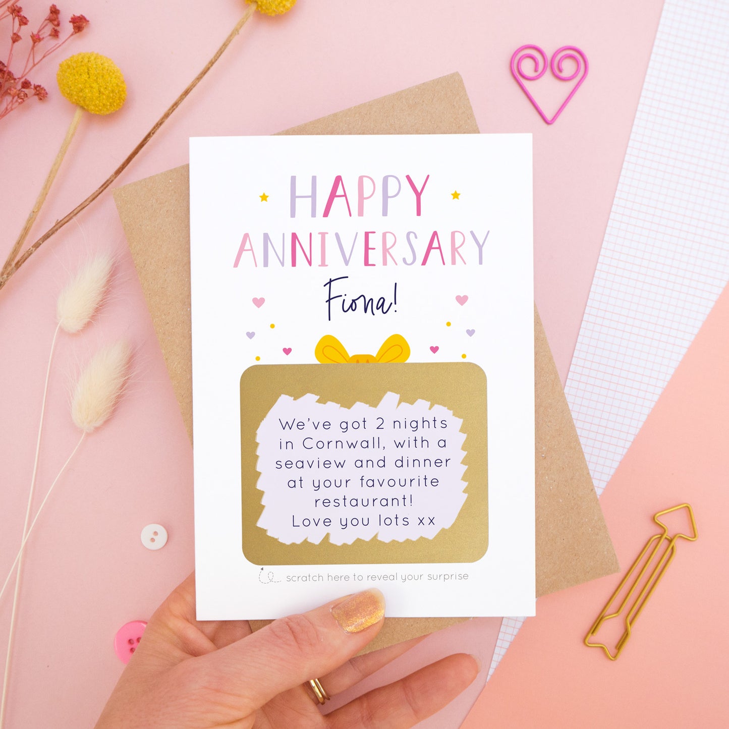 A personalised happy anniversary scratch card photographed on a pink background with floral props, paper clips, and buttons. The card has been scratched off to reveal the hidden message and is in the pink and lilac colour scheme.