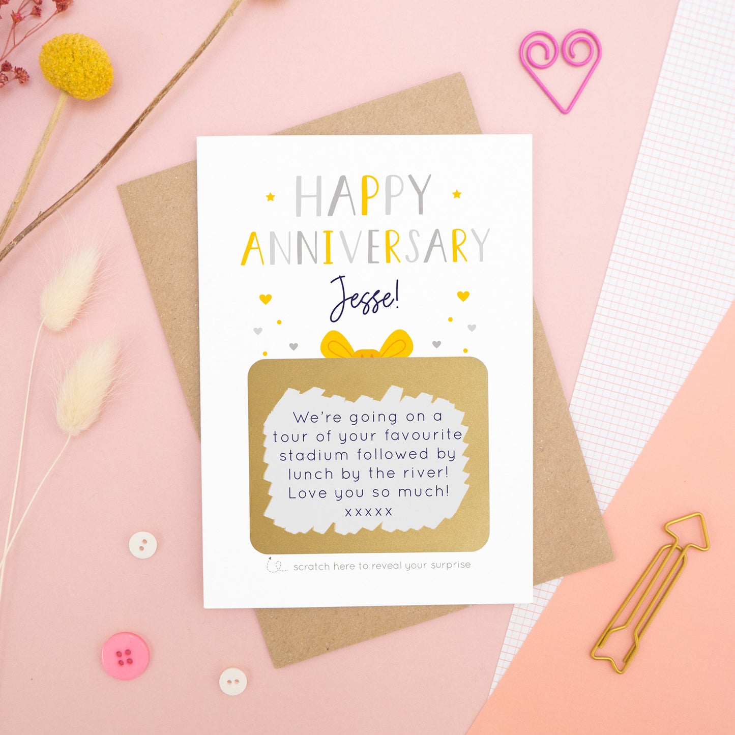 A personalised happy anniversary scratch card photographed on a pink background with floral props, paper clips, and buttons. The card has been scratched off to reveal the hidden message and is in the grey and yellow colour scheme.