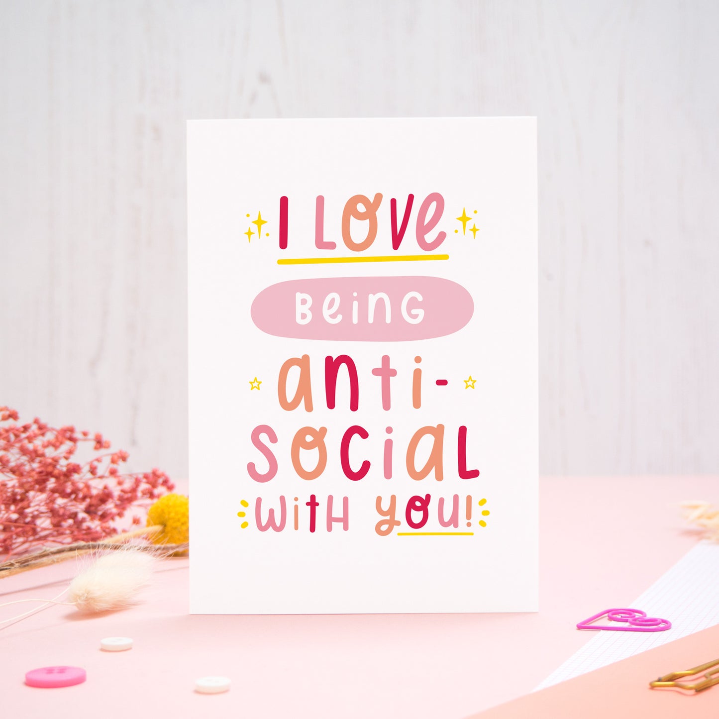 The 'I love being anti-social with you' card photographed standing up against an off white background with flowers, buttons and paperclips at the base of the card.
