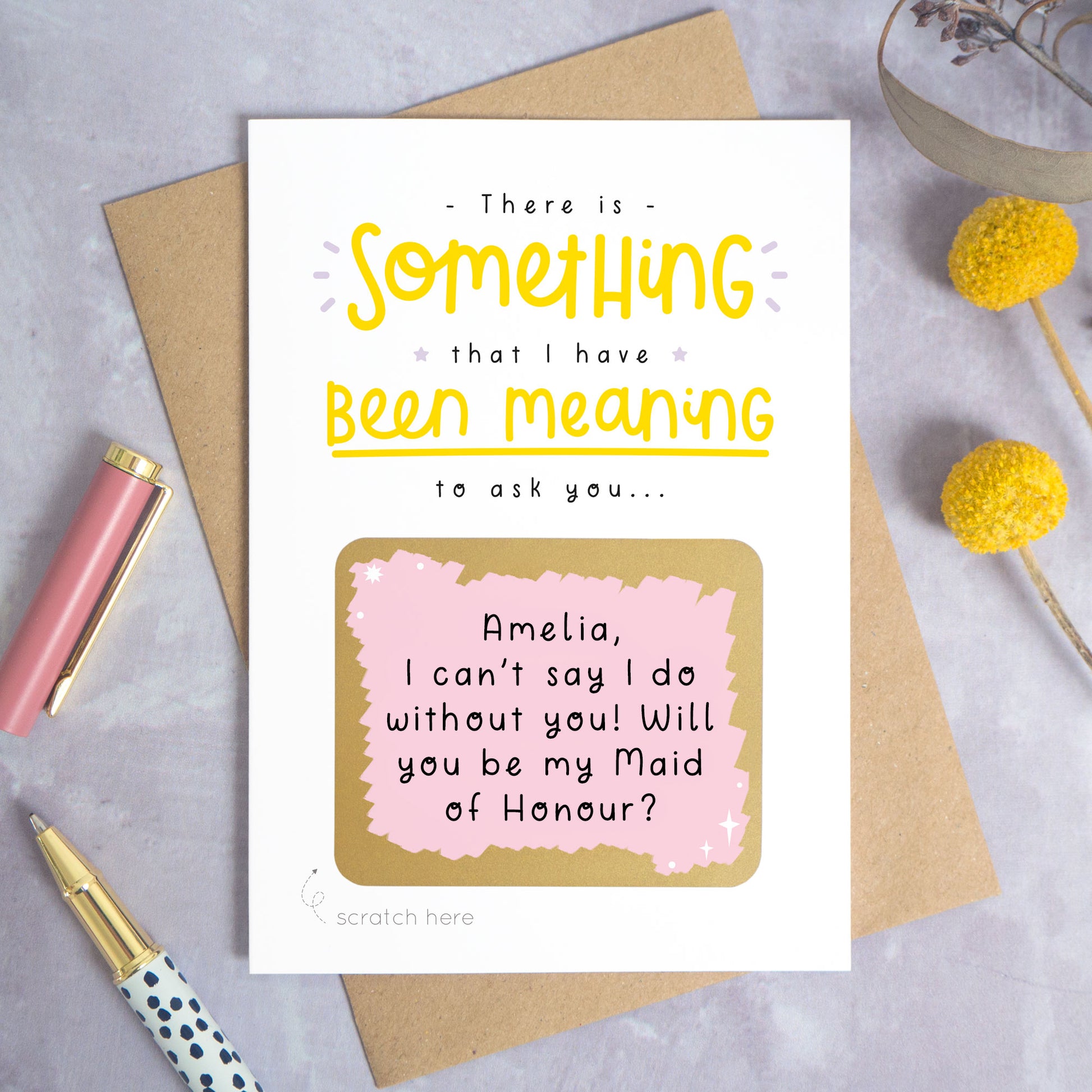 A personalised custom question scratch card photographed on a grey background with yellow flowers and a pink pen lid and spotty pen. The card is lying on a kraft brown envelope and the scratch box has been scratched off to reveal the personalised message. This is the yellow and pink colour way.