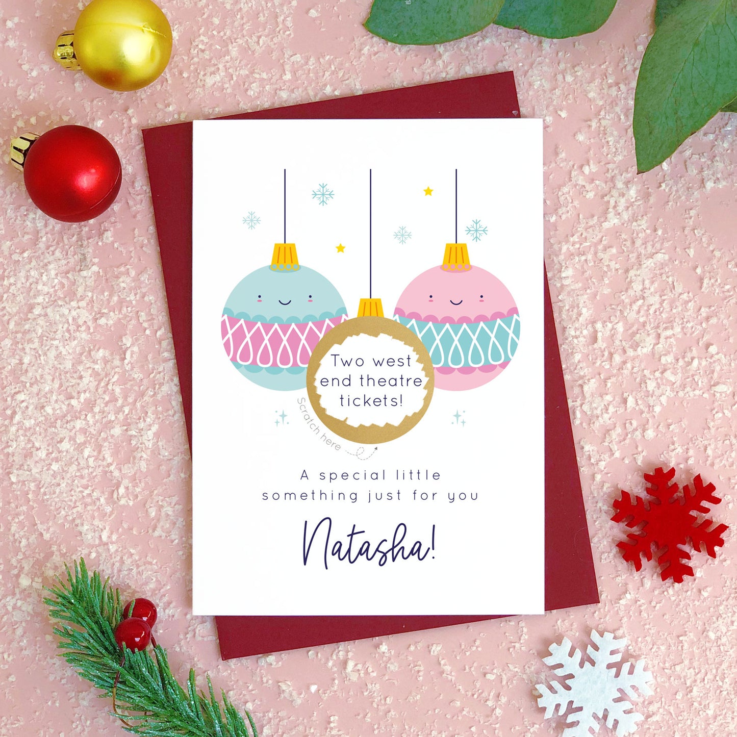 A personalised Christmas bauble scratch card photographed flat lying on a red wine coloured envelope on a pink surface surrounded by fake snow, baubles and foliage. The card is the pink and blue bauble version and the gold panel has been scratched off to reveal the custom message.