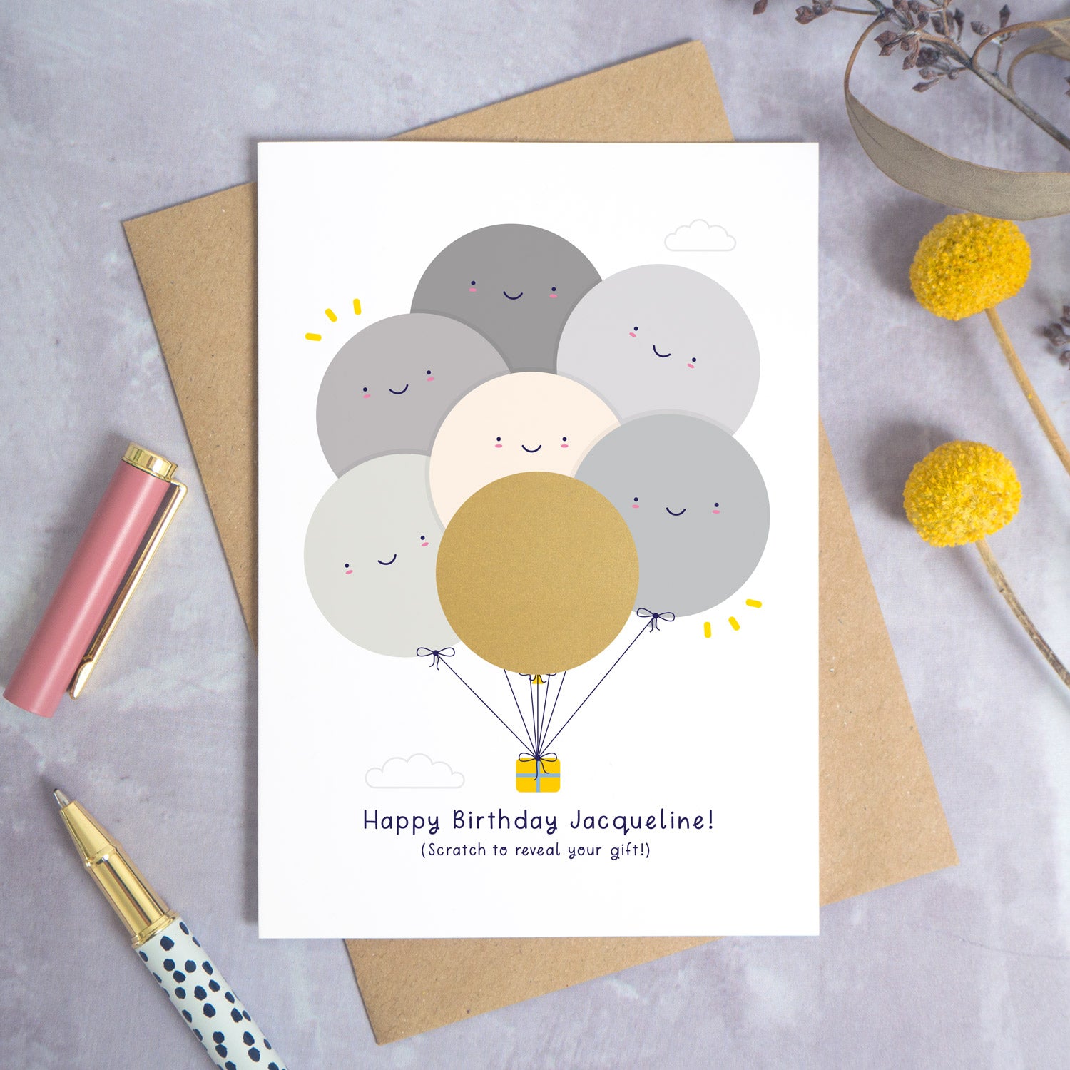 A ‘happy birthday’ scratch card photographed on a grey background with a pen on the left and yellow and purple flowers on the right. This is the grey version of the card before the gold panel has been scratched off.