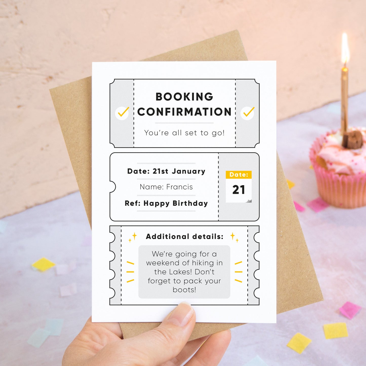 A personalised birthday booking confirmation gift card held in front of a peach and grey background scattered with birthday confetti and a cup cake with a lit candle. Pictured is the grey version of the card.