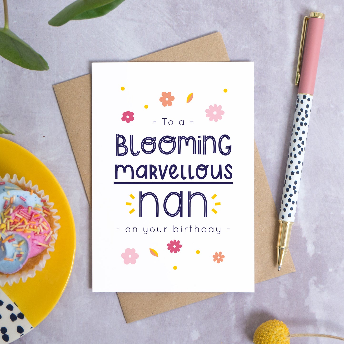 A blooming marvellous nan birthday card photographed on top of it’s kraft brown envelope set on a grey background. To the left is a yellow plate with a birthday cupcake and to the top is green foliage. To the right is a spotty pen for scale and a pop of a yellow flower in the bottom right.